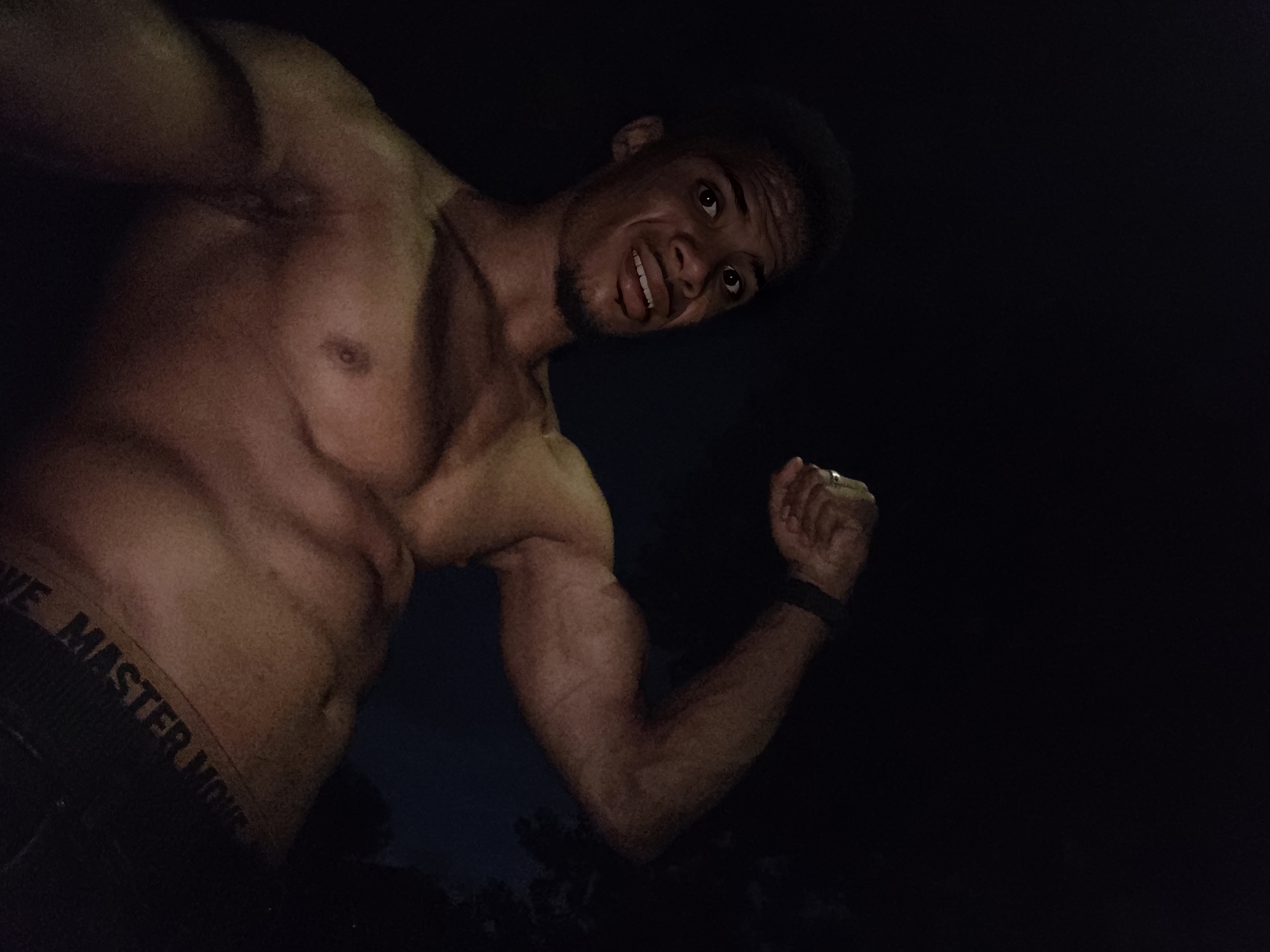 The image shows a shirtless muscular man in the dark, flexing his bicep. He is wearing black underwear with a visible logo that reads "MOVE MASTER MOVE". The man has short, dark hair, a goatee, and is looking towards the camera with a surprised expression. His skin is tan, and his muscles are well-defined. The background is dark, with some indistinct foliage visible on the right side. There is no discernible location in the city of Varna, Bulgaria.