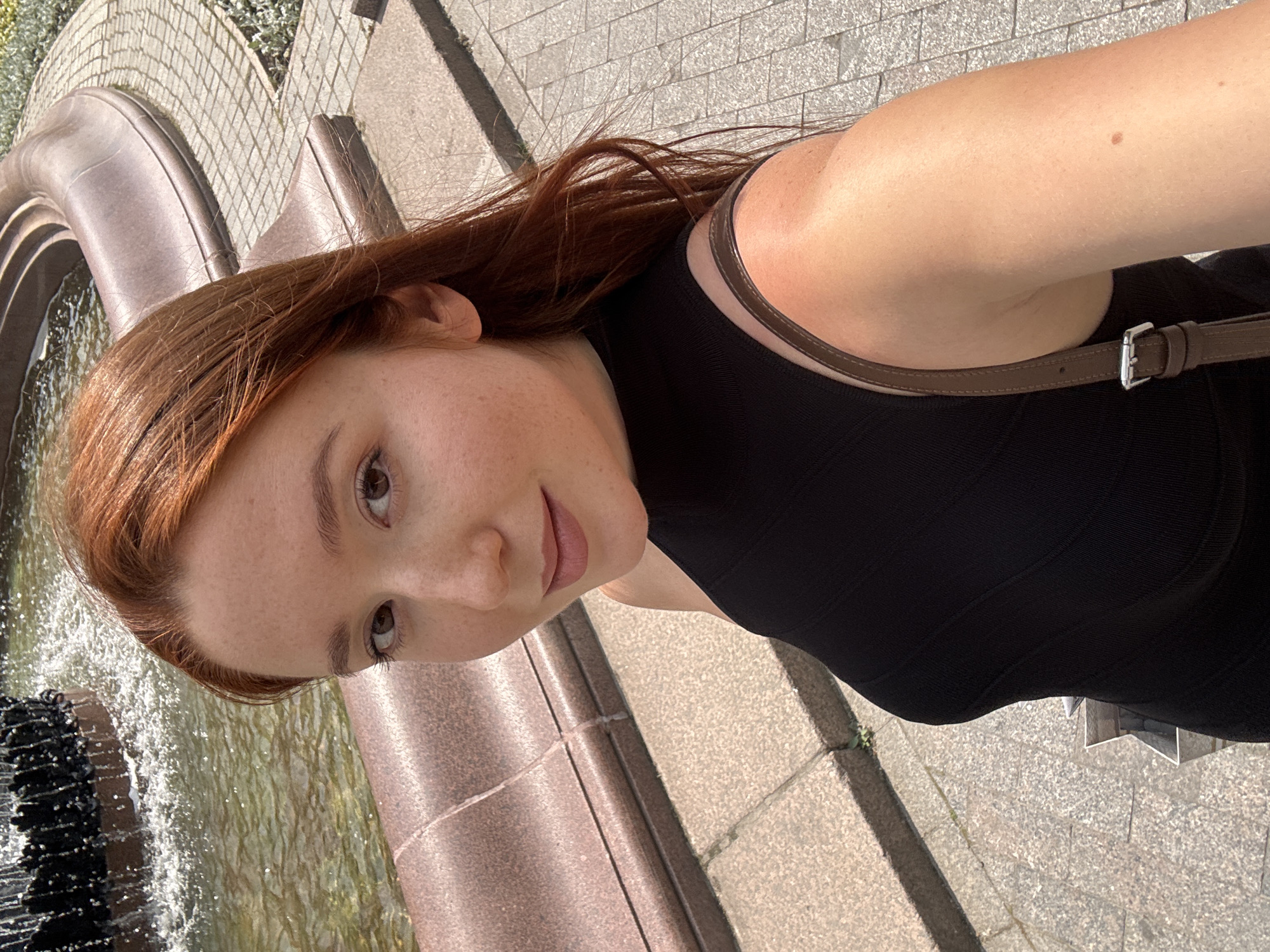 A young woman with red hair and freckles is taking a selfie outdoors. She is wearing a black sleeveless top and a brown shoulder bag. She is standing in front of a fountain with water flowing. The ground is paved with stone tiles. The scene is brightly lit, suggesting it is daytime. There are no other people visible in the frame. The overall impression is of a candid, casual moment captured on a sunny day. The location is identified as Moscow, Russia.