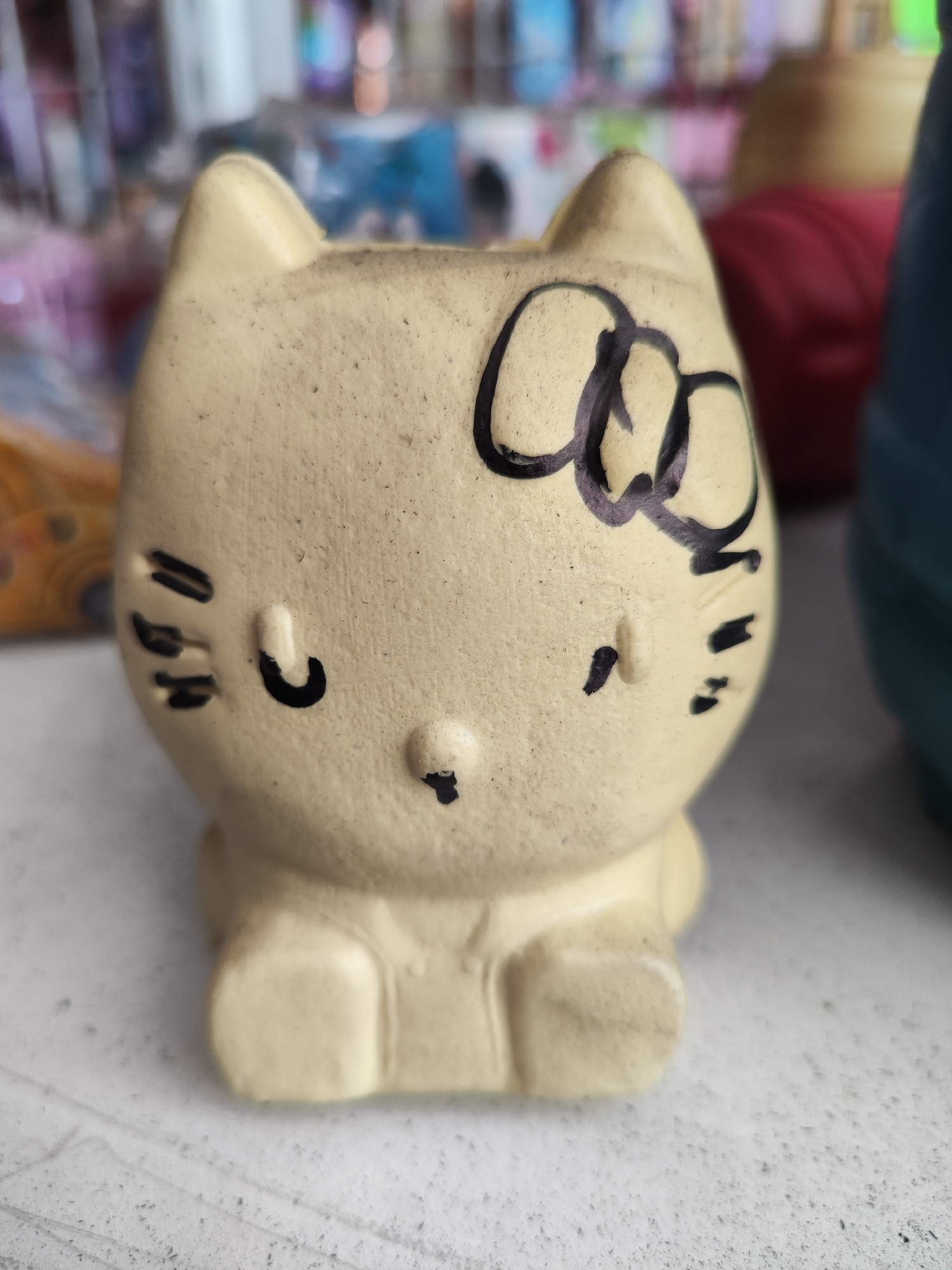 A light yellow ceramic Hello Kitty planter is centered on a light gray, textured surface. The planter has black painted details including the eyes, nose, whiskers, and a bow on its head. The background is out of focus and appears to be an indoor setting with shelves of items and some brightly colored objects. The image is taken from a slightly low angle, with the planter filling most of the frame.