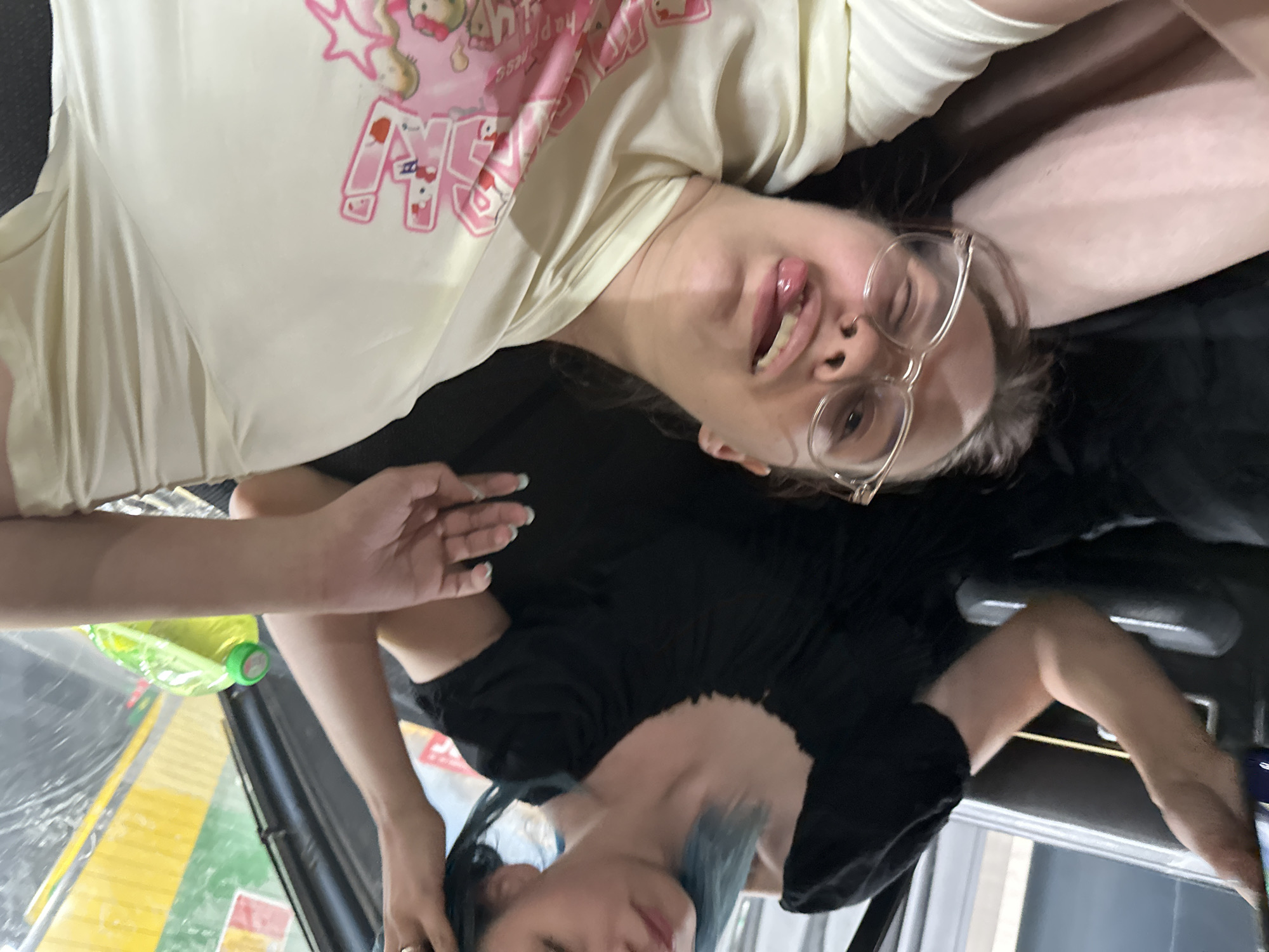 This image shows two people in what appears to be a casual setting, possibly a vehicle. One person is wearing a light-colored t-shirt with a graphic print featuring pink characters and text. This individual is looking towards the camera with their tongue sticking out slightly and wearing clear-rimmed glasses. The other person, mostly obscured, is wearing a black t-shirt. Their hair has streaks of blue.
A bright green plastic bottle with a green cap is visible in the foreground, resting on a surface that appears to be a car's interior. The background is dark and out of focus. The lighting suggests it is daytime, and no specific weather conditions are discernible. The overall impression is one of a candid, informal moment.
The text on the t-shirt appears to include some stylized letters, possibly "M" and "NG," along with some smaller, less legible words that could be English. The graphic on the shirt also includes star shapes and cartoonish characters, which might be related to anime or a similar style. The scene is likely indoors or in a shaded area, given the lighting and reflections.