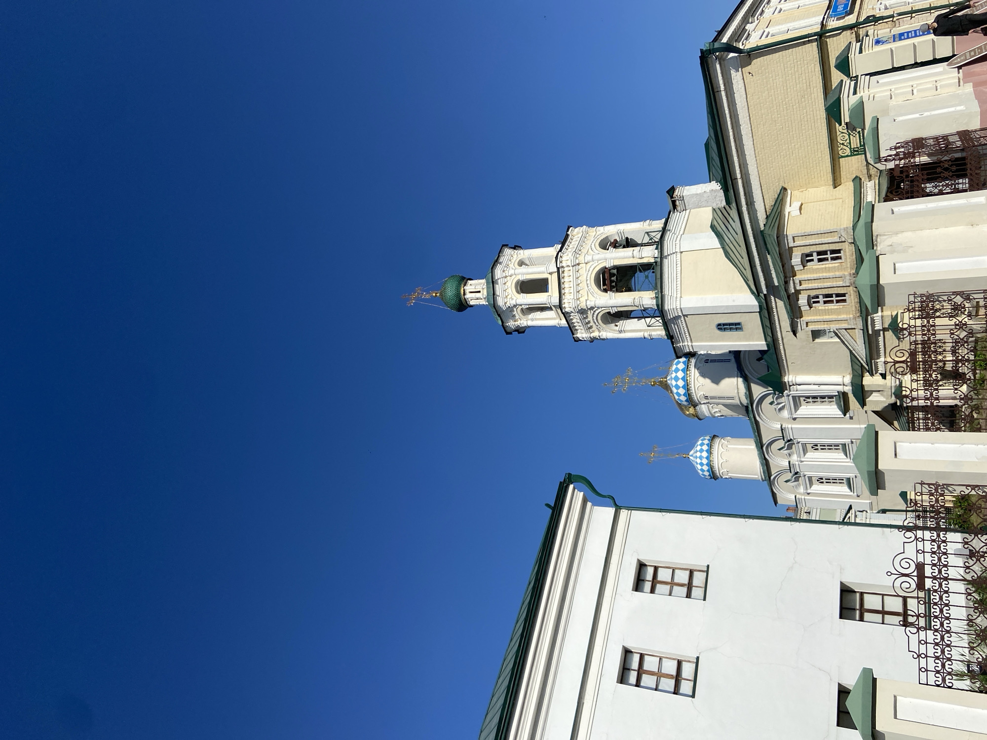 The image captures a low-angle view of the Annunciation Cathedral and its bell tower within the Kazan Kremlin, Kazan, Russia. A bright blue sky fills much of the frame. The architecture includes white and yellow buildings with elements of green trim and ornate ironwork. Distinctive blue and white domes with golden crosses are visible, as is the white bell tower featuring green-domed tops. The architecture also displays a mix of baroque and classical styles. A street sign is visible in the top right.