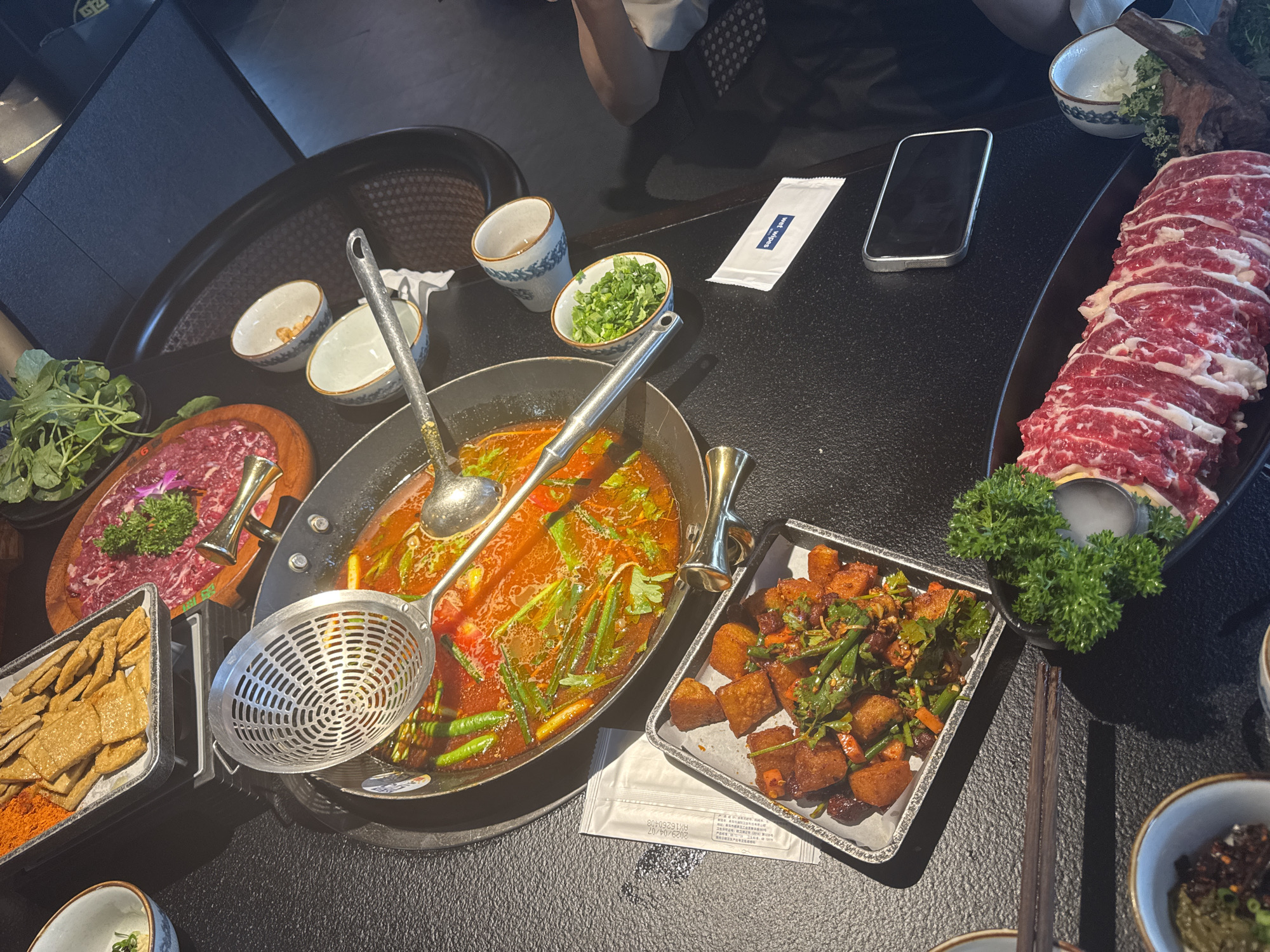 The image captures a tabletop arrangement of various dishes in what appears to be a restaurant setting. A hot pot filled with a red-tinted broth and green vegetables sits in the center, accompanied by a ladle and a straining skimmer. Surrounding the hot pot are several plates and trays containing different ingredients.
To the left of the hot pot, there's a wooden board with thinly sliced meat, garnished with fresh greens and what looks like edible flowers. Adjacent to it, there's a tray of rectangular fried snacks, possibly tofu or bean curd, dusted with reddish seasoning.
To the right, a tray filled with bite-sized fried foods mixed with green vegetables and red chili flakes can be seen. A platter of thinly sliced raw beef, arranged in a circular pattern, dominates the upper right corner, decorated with a small bundle of leafy greens.
Small bowls, some containing sauces or condiments, are scattered around the table. A smartphone rests near the platter of beef, and a white packet, possibly containing wet wipes, is also visible. Part of a person can be seen in the upper portion of the image.
Given the prevalence of hot pot and the style of the dishes, this photograph might have been taken in a restaurant specializing in Sichuan cuisine, which is known for its spicy and flavorful dishes. The exact location within Yuzhong District, China, cannot be determined solely from this image, as the context clues are only based on the style of food and dining arrangement.