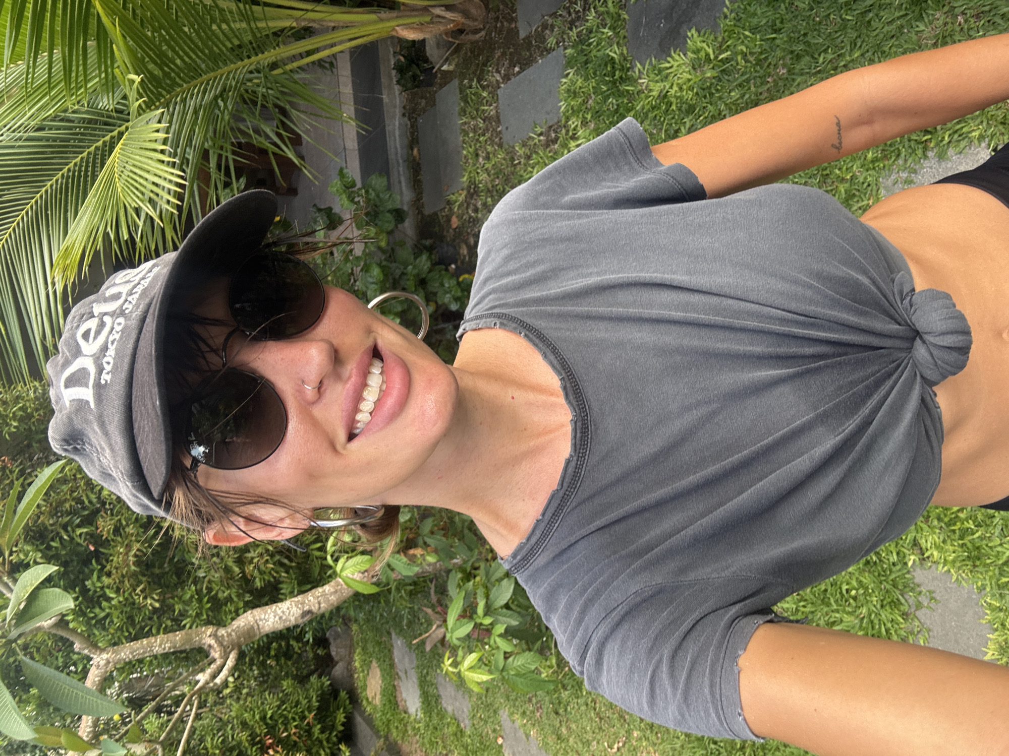 A woman is seen in a full-body selfie taken in North Kuta, Indonesia. She is wearing a gray T-shirt knotted at the front, black bottoms, a baseball cap, and sunglasses. She is smiling and has a nose ring and hoop earrings. The background consists of green foliage, including palm trees and other plants, with grass and stone tiles visible on the ground.