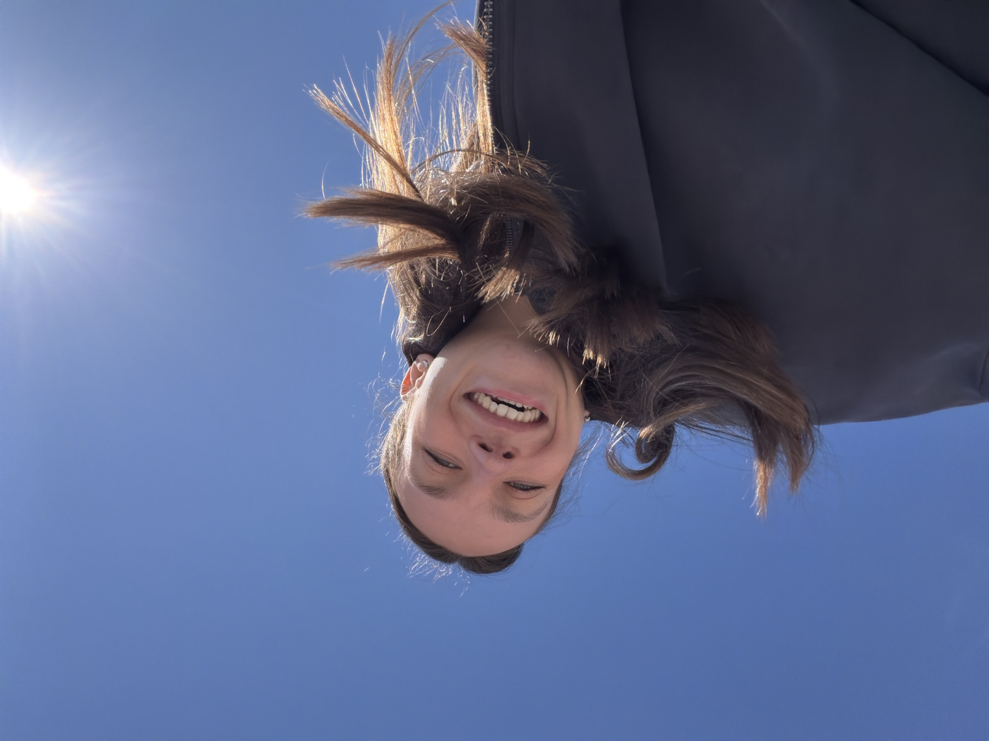 A woman smiles at the camera from an upside-down perspective. She has fair skin, and her brown hair is illuminated by the sun to the upper left of the frame. She is wearing a dark-colored jacket. The background is a clear blue sky with a bright sun visible. The location is not specified in the image.