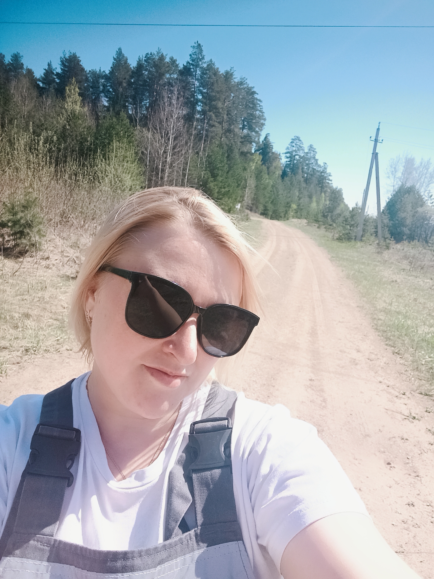 A selfie taken in a natural environment on a sunny day. A fair-skinned person with blonde hair and wearing sunglasses, a white t-shirt, and gray overalls is in the foreground. Behind the person is a dirt road leading into a forest. Power lines and poles are visible on the right side of the road. The location appears to be near a forest in Chaikovsky, Russia.