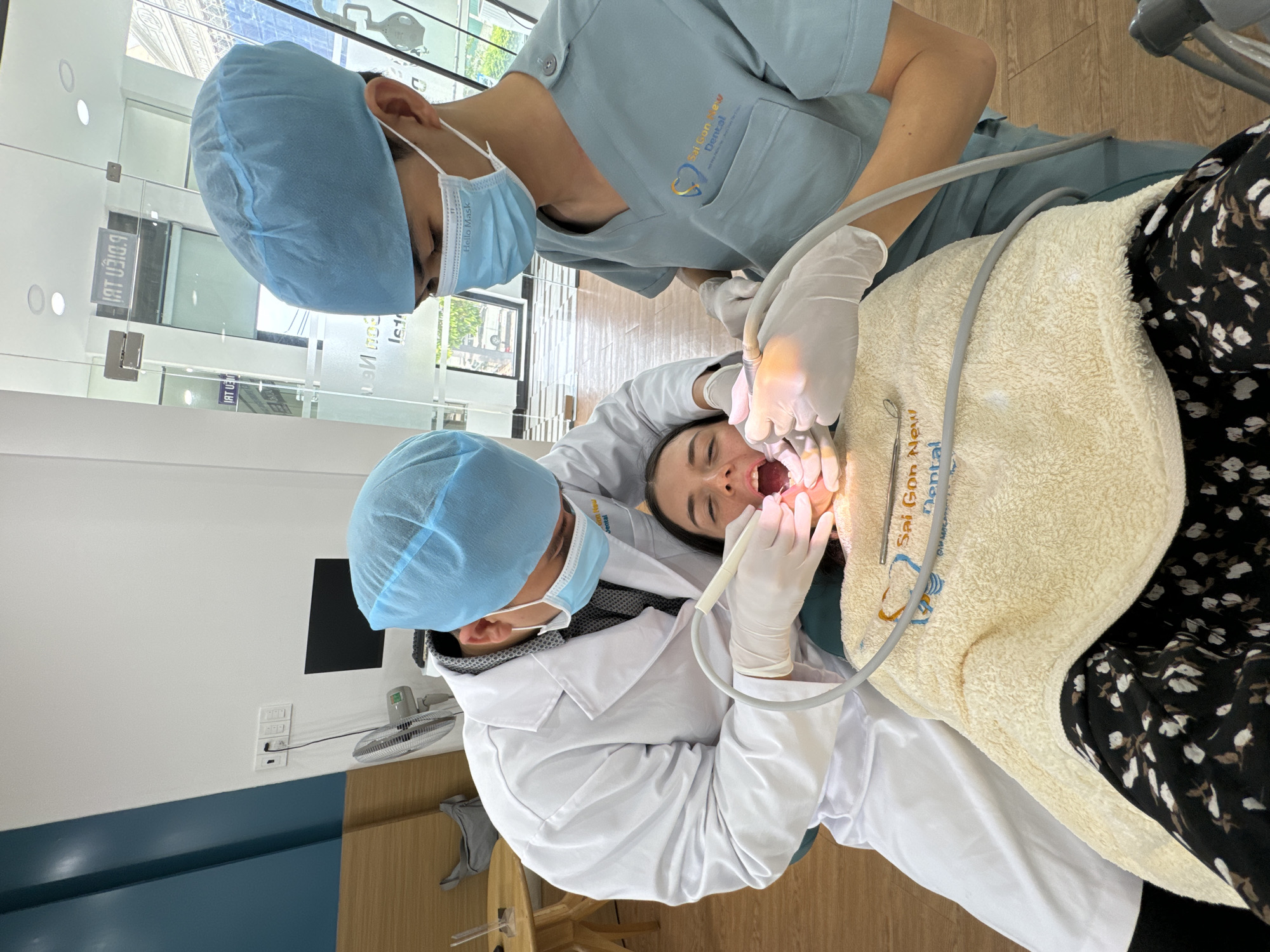 A horizontal, eye-level shot captures a dental procedure taking place in a modern and brightly lit dental clinic, likely in Phu Thuy Ward, Vietnam. A female patient is reclined in a dental chair, her mouth open for treatment. She is covered by a light-colored, textured towel or blanket, which displays the blue "Sai Gon New Dental" logo. She wears a dark dress with a white floral pattern.
Two dental professionals are attending to her. One, wearing a white lab coat, light blue surgical cap, and white gloves, holds a dental tool with a light, performing the procedure inside the patient's mouth. This professional's face mask has "Hello Mask" printed on it. The second professional, wearing light blue scrubs with the "Sai Gon New Dental" logo on the pocket, a matching surgical cap, and a face mask, stands slightly behind, appearing to assist or observe.
The background shows a clean environment with white walls, light-colored wooden flooring, and a glass partition. Mirrored text is visible on the glass, including "SDIEN IBI" and "ONG GROUPH," along with "Sai Gon New Dental". A small electric fan and power outlets are visible on a wall to the left. The scene depicts a focused and professional dental examination or treatment.