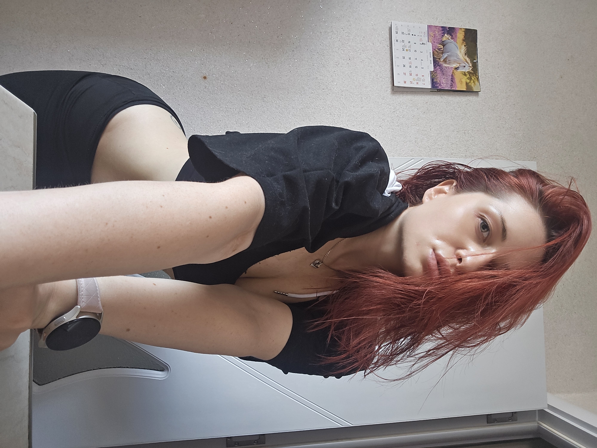 An indoor, horizontally oriented medium shot depicts a young woman with long, reddish-brown hair, looking directly at the viewer with blue/gray eyes. She is positioned as if lying down or propped up, with her head slightly tilted and her hair splayed out. Her expression is neutral to slightly pensive. She is wearing a black v-neck top or dress, with a white strap (possibly a bra strap) visible on her chest, along with a gold-colored necklace featuring a small pendant.
On her left wrist, she wears a silver-colored smartwatch with a light pink, textured band. Her skin is fair, showing visible freckles or moles on her arms. A faint, dark tattoo is discernible on her upper right thigh or hip area, partially covered by her black clothing. The black fabric of her top appears to have some light lint or dust.
The background features a plain, light-colored, slightly textured wall. In the upper right quadrant, a wall calendar for "ЮНИ 2026" (June 2026) is visible, displaying dates including 3, 4, 5, 6, 7, 10, 11, 12, 13, 15, 24, 25, 26, 27, and 2, 3, 4, 5. The calendar features an image of a white horse running through a field of lavender. To the woman's right, a white door or cabinet front is visible. On her left, a light-colored, possibly marble-patterned surface (such as a table edge) is partially in view. The scene takes place indoors, likely in a residence in Burgas, Bulgaria. The lighting is soft, indicative of an interior setting.