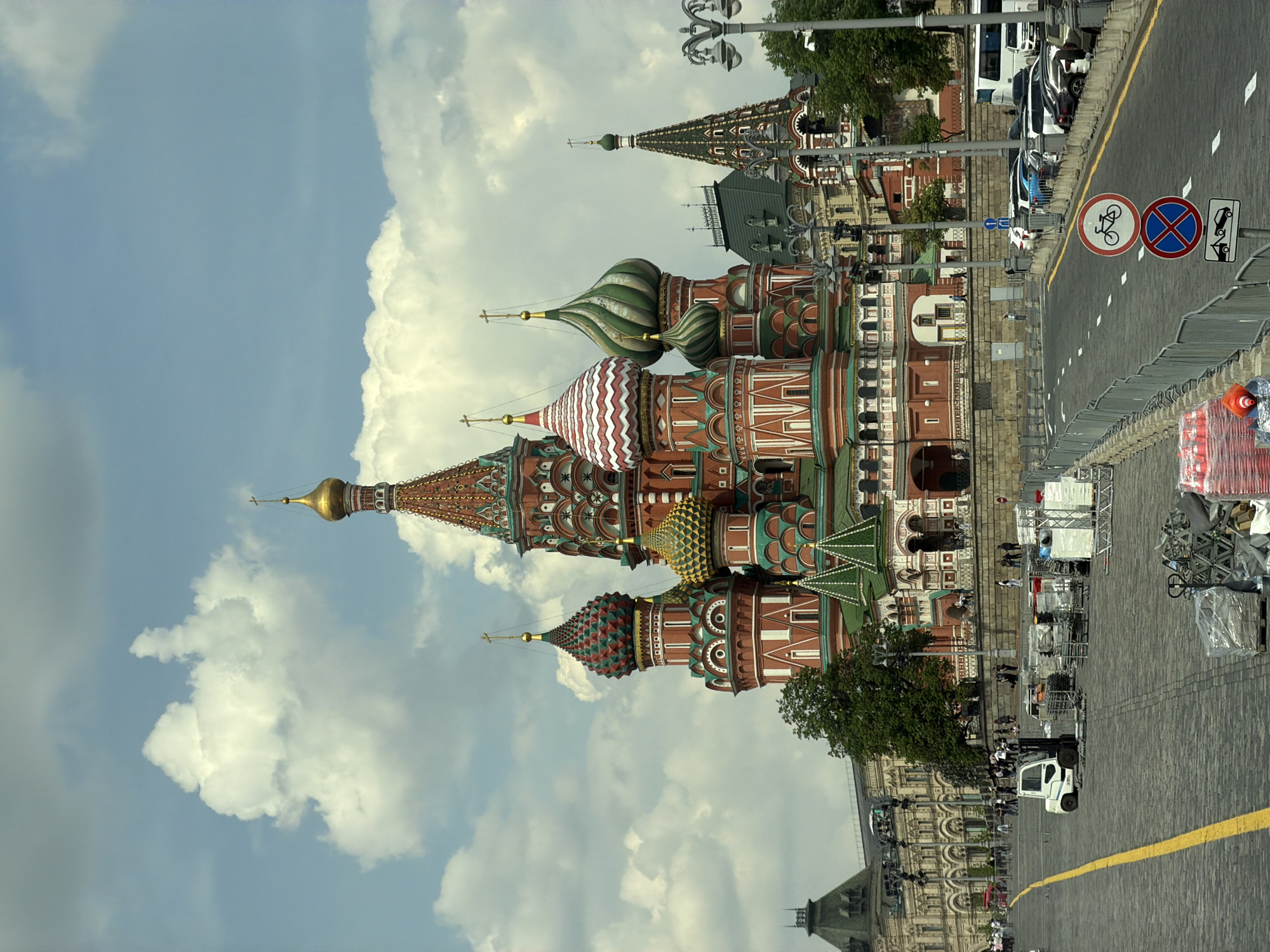 This image shows the iconic Saint Basil's Cathedral in Moscow, Russia, against a backdrop of partly cloudy sky. The cathedral, with its distinctive colorful onion domes, stands next to a pedestrianized area. The photo also captures a street with a yellow line and a "no parking" sign, indicating an urban environment with traffic regulations. Adjacent to the street, there are metal barriers, possibly for crowd control or event preparation. Construction or event setup is also suggested by the presence of scaffolding and equipment alongside the pedestrian area. There are several people moving around the scene.