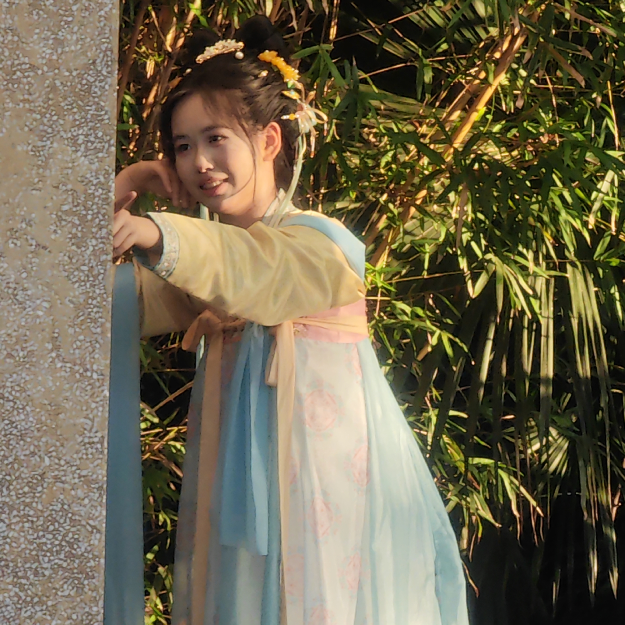 A young woman stands next to a patterned column. She is wearing a traditional East Asian dress in pastel colors (yellow, blue, and pink) with floral designs and has decorative hair ornaments. The background consists of green bamboo stalks. The scene appears to be in a garden or park setting, potentially within Yuzhong District, China. The woman's pose suggests she is looking towards something or someone out of the frame.