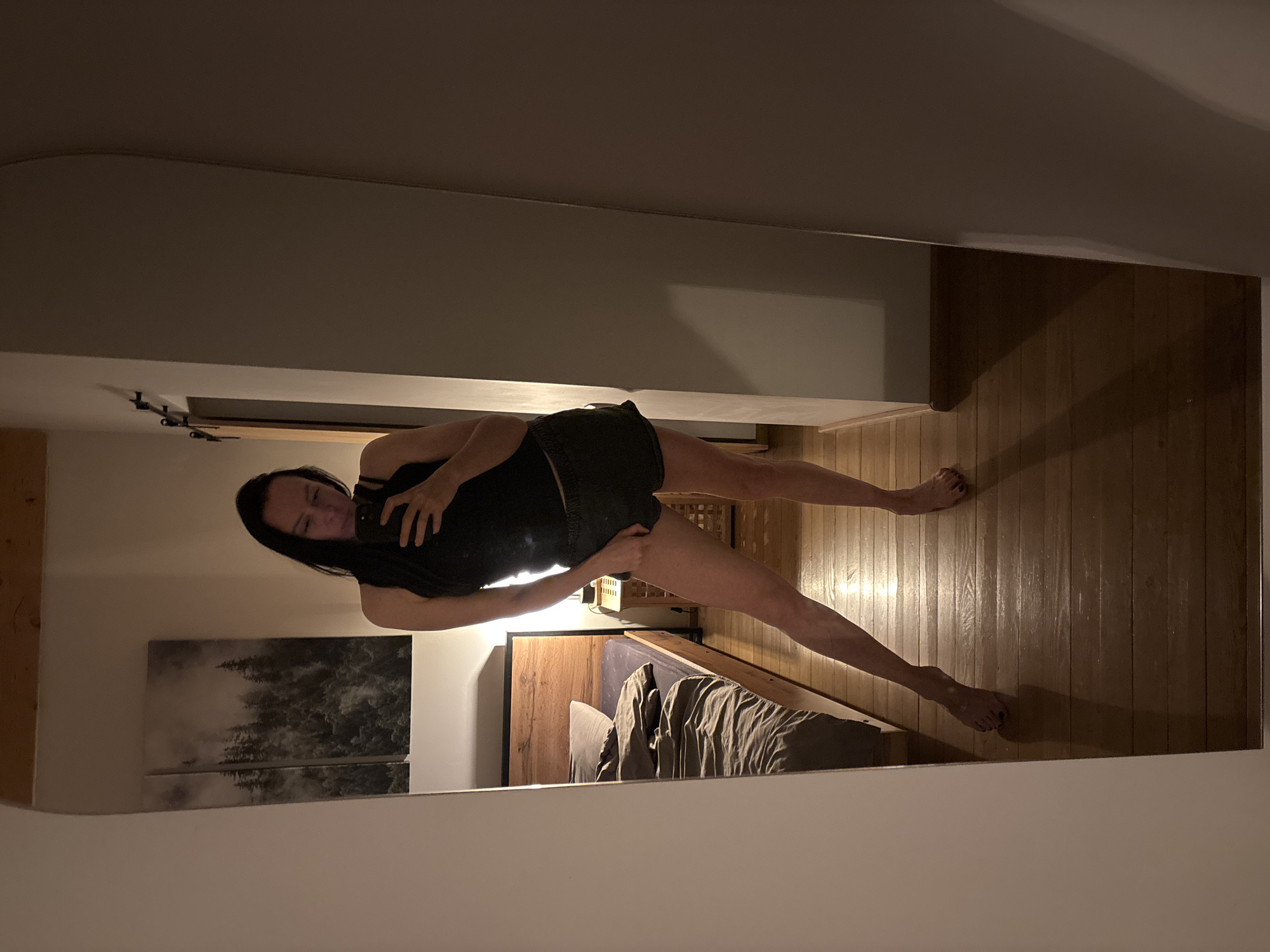A mirror reflects an image of a woman standing in what appears to be a bedroom. She is holding a smartphone in her right hand, seemingly taking a selfie. She is wearing a dark top and shorts. The bedroom features hardwood flooring, a bed with brown bedding, a wooden headboard, and a piece of artwork depicting trees. The location cannot be determined as Chaikovsky, Russia, based on the visual information provided.