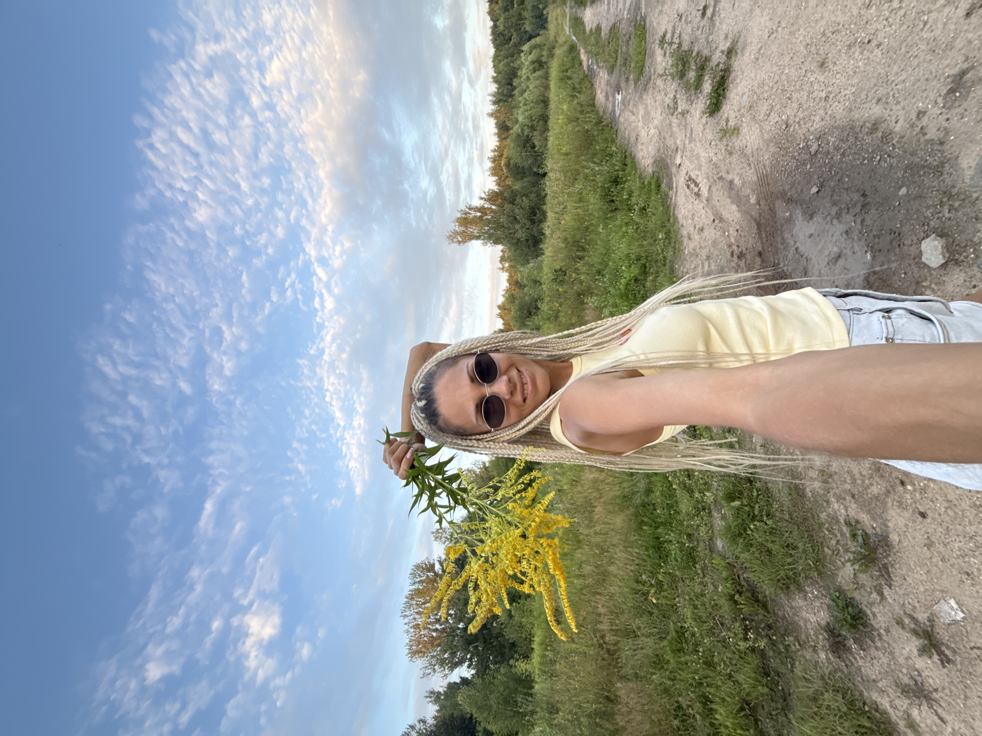 A light-skinned woman with blonde braided hair and sunglasses holds a yellow bouquet of flowers above her head while smiling at the camera. She wears a light yellow tank top and denim shorts. The woman stands on a dirt path in a grassy, wooded area with a partly cloudy sky in the background. The image appears to be taken in Yaroslavl, Russia.