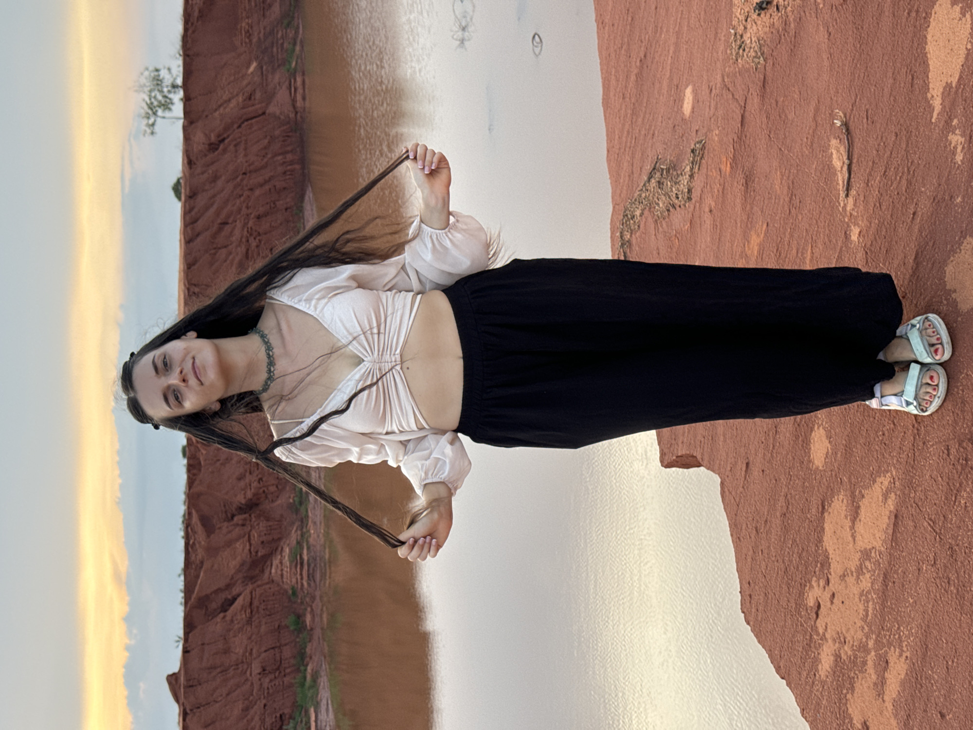 A young woman stands on a reddish-orange cliffside overlooking a calm body of water, likely in Mui Ne, Vietnam. She is positioned towards the left, looking directly at the camera with a gentle smile. She has long, dark hair and is holding strands of it in front of her with both hands.
She wears a light, off-white, long-sleeved crop top with a ruched front that reveals her midriff, paired with black wide-leg trousers. On her feet, she has light-colored open-toed sandals, with the word "HELLO" visible on the strap of her right sandal. Her toenails are painted red. A dark, possibly beaded, choker necklace is visible around her neck.
The landscape features distinct red-orange earth, possibly clay or sandstone formations, with some textured patterns and sparse dry vegetation on the ground she stands on. The water to her right is placid, reflecting the soft light. In the background, across the water, another similar reddish-orange bank or cliff is visible, topped with a small, sparse tree. The sky above shows warm, muted tones, suggesting the scene takes place during late afternoon or early evening. The weather appears clear and calm.