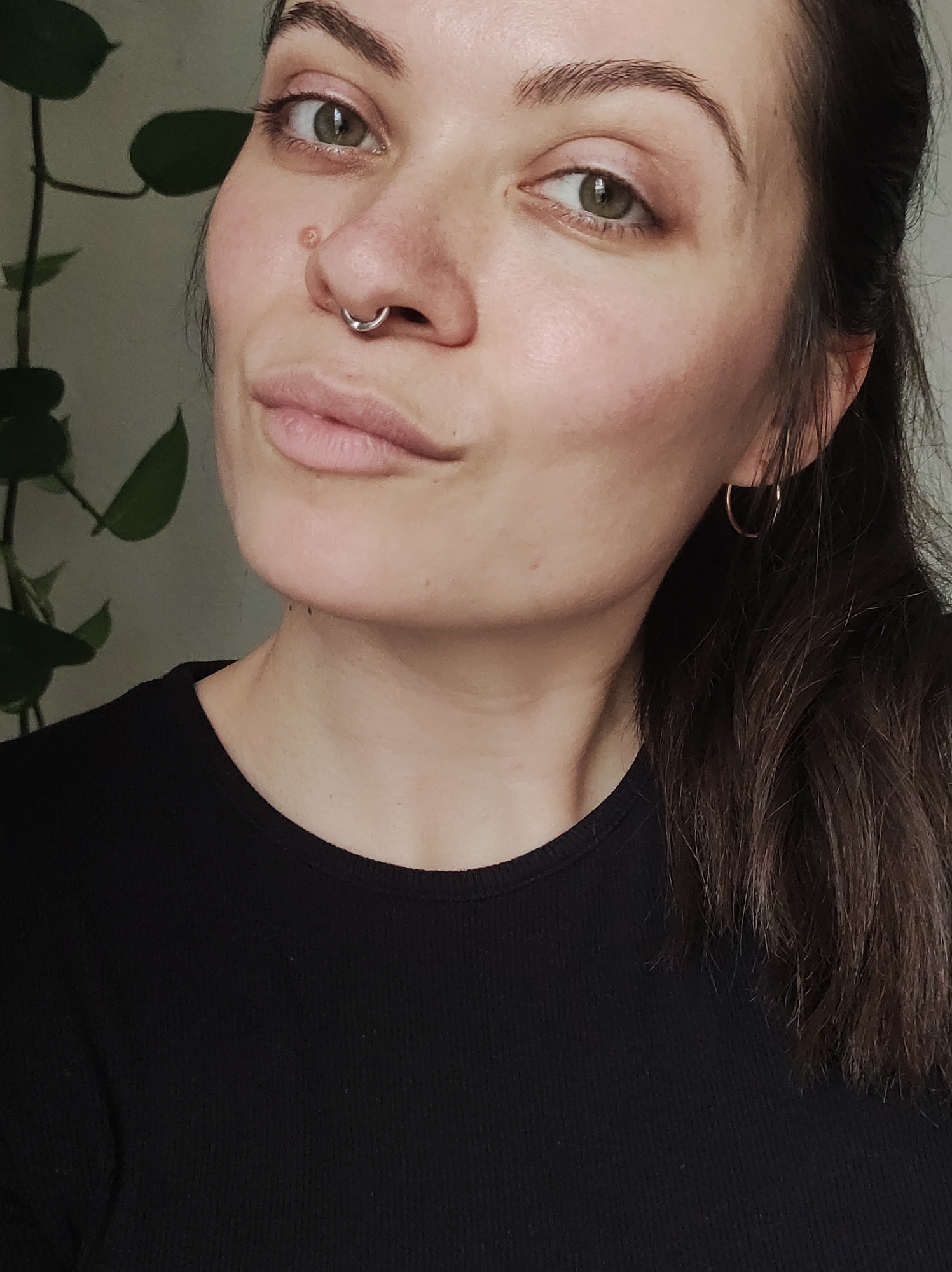 A young woman with light skin and brown hair pulled back from her face poses for a close-up shot. She has hazel eyes and is wearing a silver septum ring in her nose. A small mole is visible near her nose. She is also wearing small gold hoop earrings. She is wearing a black, long-sleeved shirt. Part of a green plant is visible in the background on the left side of the frame. The woman's expression is slightly pensive, with a subtle smile. The background is a light color. There is nothing to indicate the location is Chaikovsky, Russia.