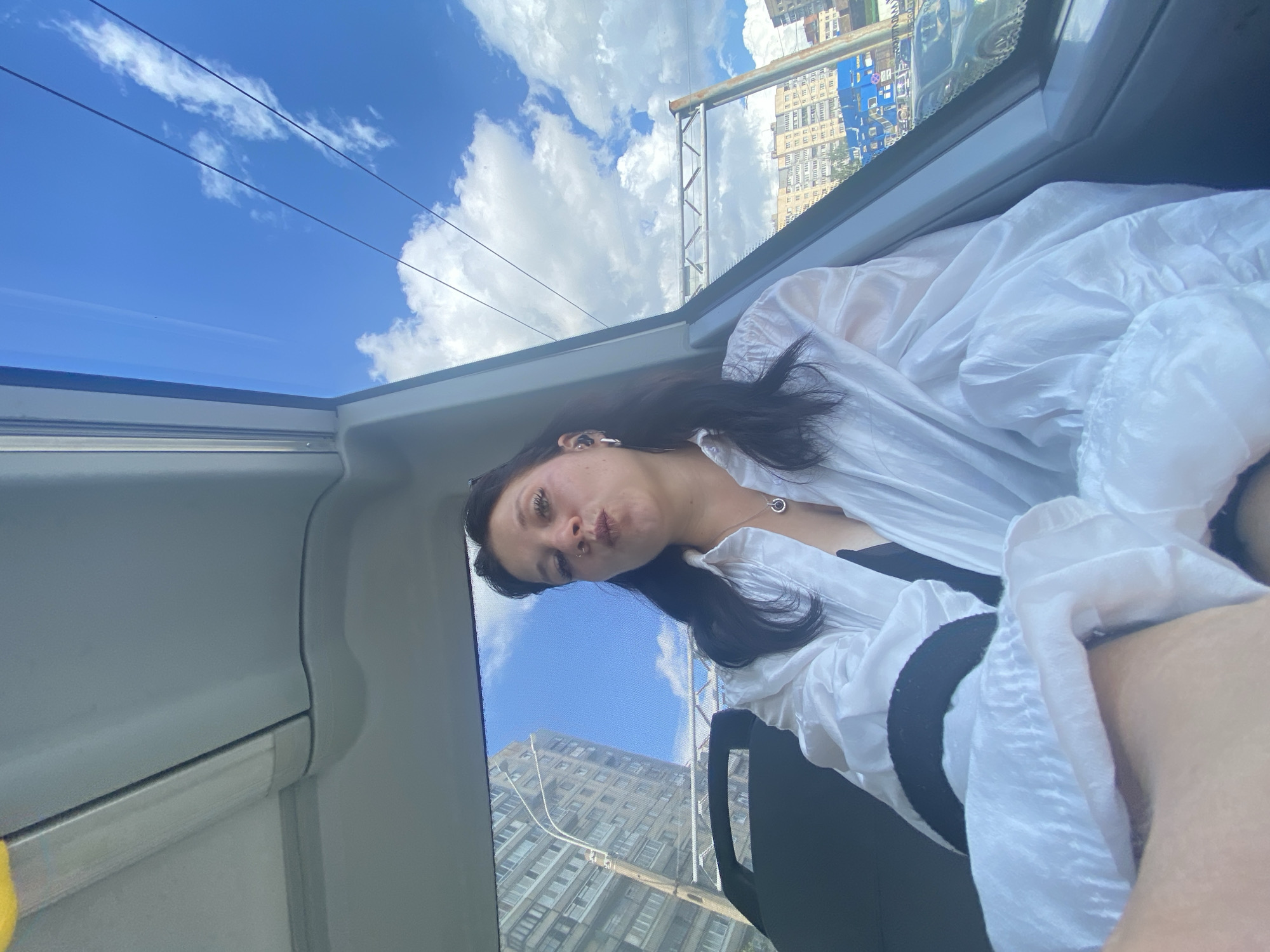 In this image, a woman with dark hair and light-colored clothing sits inside what appears to be public transportation. She is facing the camera, with her mouth slightly pursed. She is wearing a white, loose-fitting shirt, a black bra or camisole underneath, and a necklace with a circular pendant. She also has earrings in her ears and a nose ring. The background shows a window with a view of city buildings, a blue sky with clouds, and overhead wires. The interior of the vehicle is visible, with grey seating and panels. The urban landscape outside suggests a city environment, likely within Saint Petersburg, Russia, given the architecture and the presence of overhead electric wires typical of the city's tram or trolleybus system.
