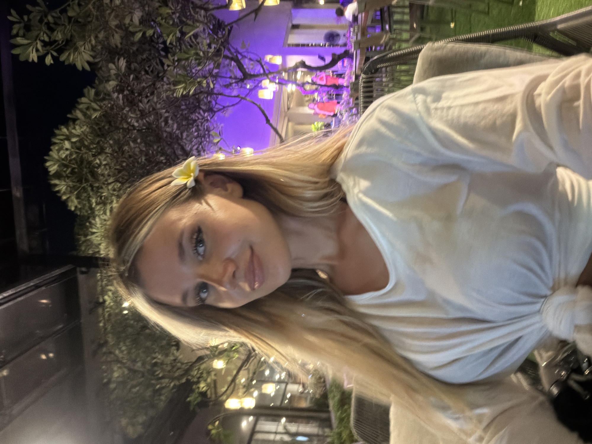 A blonde woman with a plumeria flower tucked behind her ear is seated at an outdoor restaurant in Patong, Thailand. She is wearing a white t-shirt tied at the waist. The background features lush trees, ambient lighting from lamps, and a stage with performers illuminated by purple lights. The setting appears to be a lively evening venue.