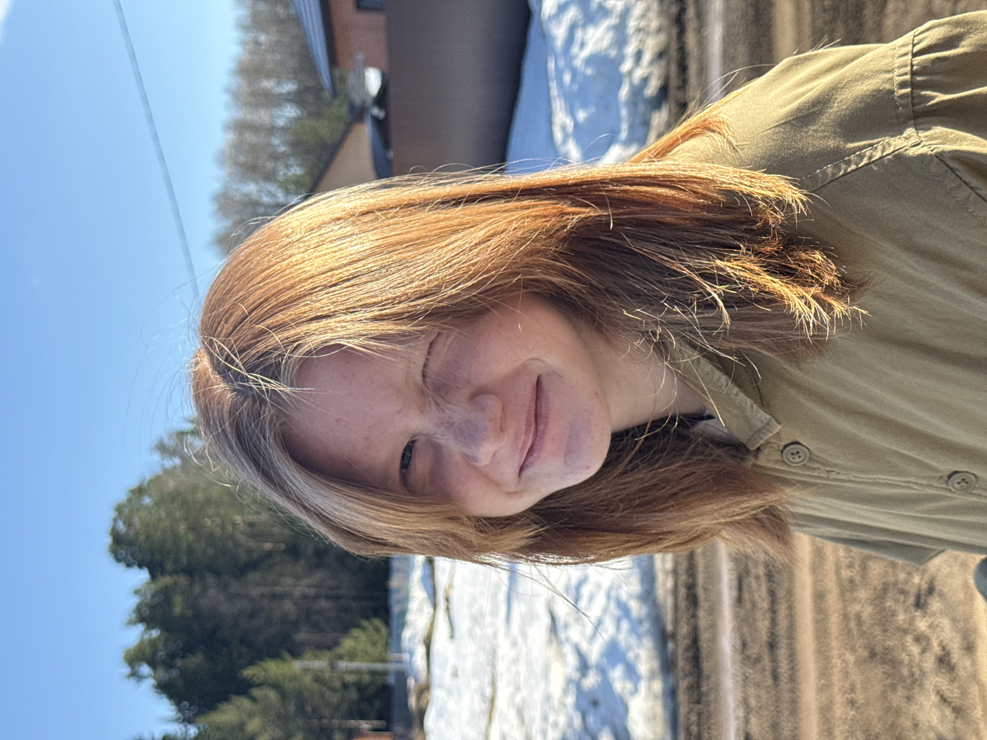 A person with light skin, freckles, and red hair, wearing a light brown button-down shirt, is standing outdoors. They are smiling with one eye closed. Patches of snow are visible on the ground in the background, along with trees and a building. The location is not definitively identifiable as Chaikovsky, Russia.