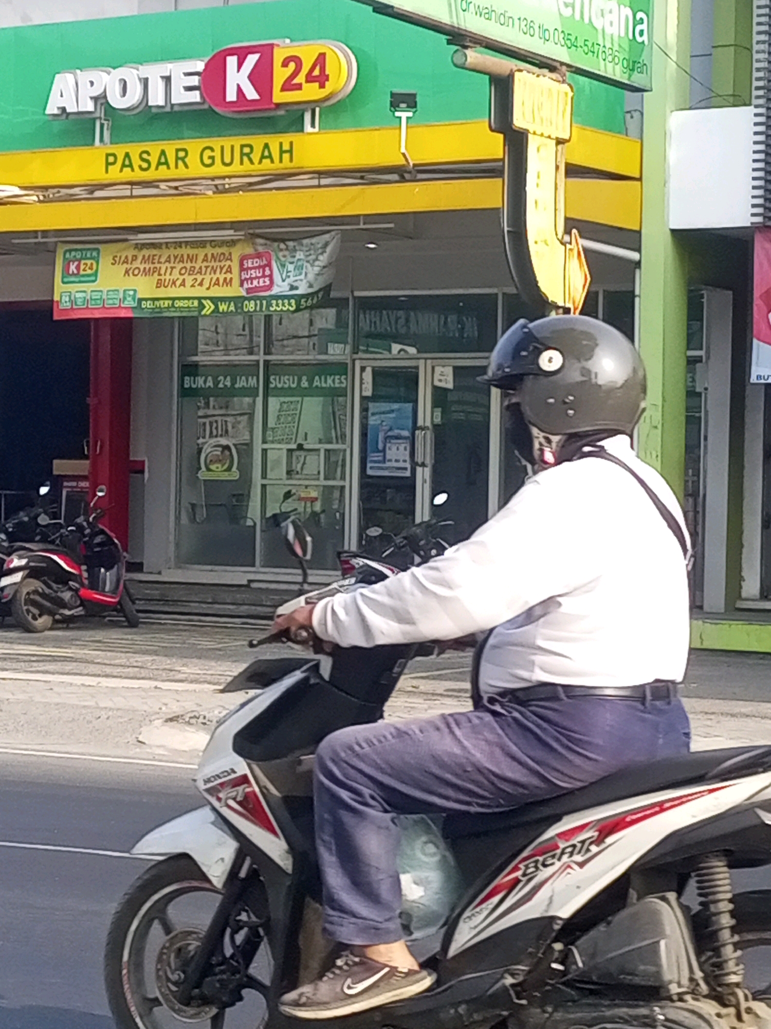 In Gurah, Indonesia, a person wearing a white shirt, blue pants, and a dark helmet rides a white and red Honda Beat motorcycle. The motorcycle has "BEAT" written on its side. The person is in front of an "APOTEK K 24" pharmacy, which has a sign reading "PASAR GURAH". A banner on the pharmacy states "SIAP MELAYANI ANDA KOMPLIT OBATNYA BUKA 24 JAM". The scene is outdoors, with a paved road and sidewalk visible. Other motorcycles are parked in the background.