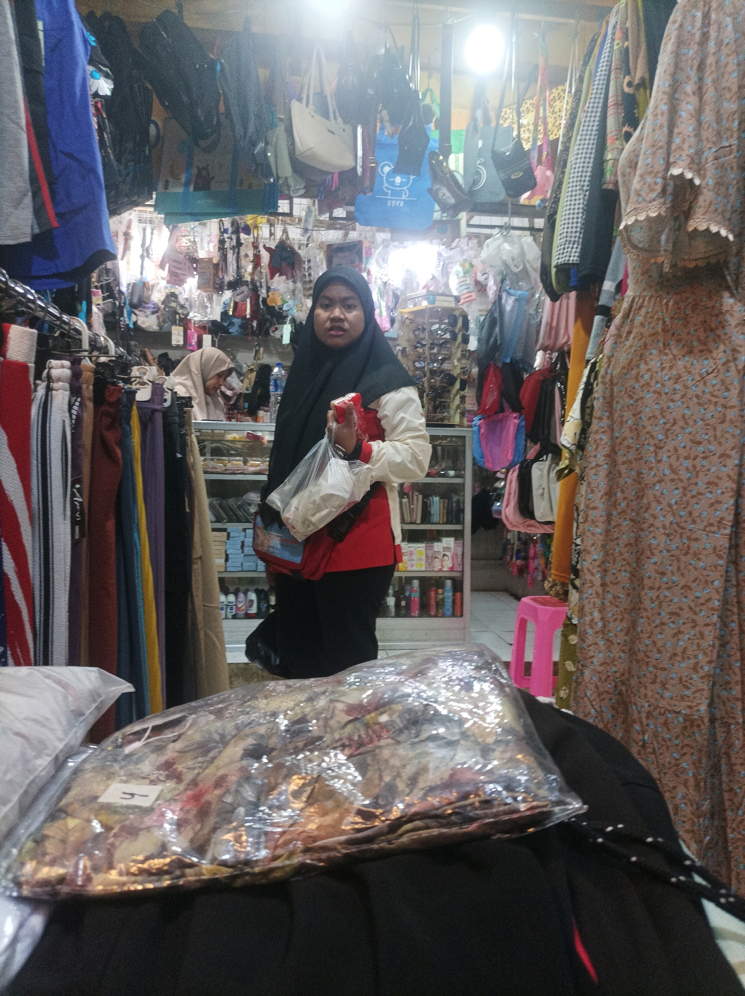 The image depicts the interior of a small clothing shop in Sumuragung, Indonesia. A woman wearing a black hijab and a white and red jacket is standing in the center of the frame, holding a plastic bag with items inside. She is looking towards the camera. In the foreground, a stack of clothing is visible, wrapped in clear plastic. The shop appears to be filled with various apparel, including dresses, pants, and bags hanging from racks. Shelves behind the woman are stocked with merchandise. The lighting is bright, suggesting an indoor commercial setting.