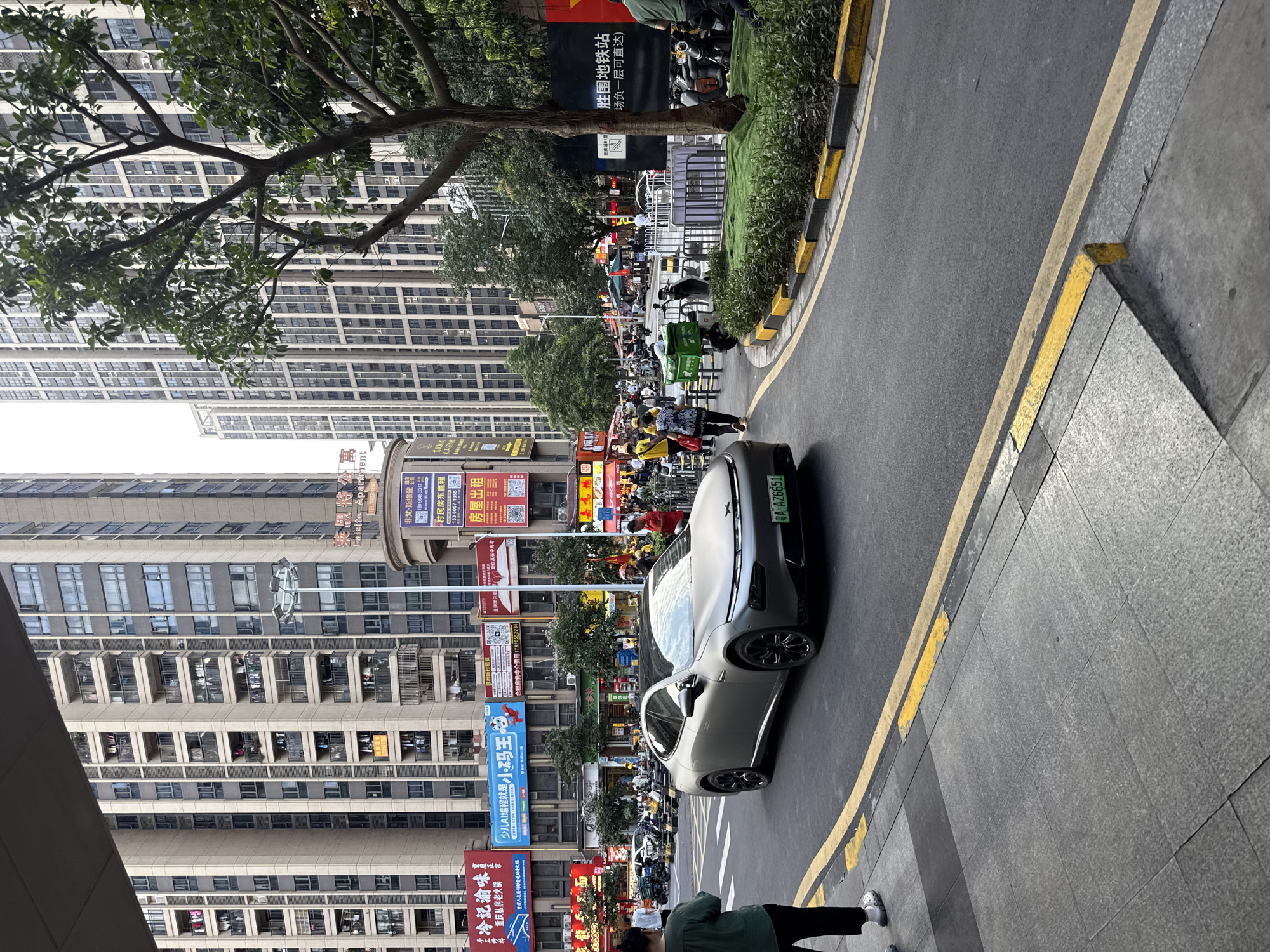 This image captures a street scene in Haizhu District, China, likely during daytime given the lighting. In the foreground, a modern, silver electric car with a black roof and black wheels is the primary subject, driving on a road marked with yellow and black curbs. The car's license plate is visible and reads "A AZ6651". A pedestrian, seen from behind and wearing dark pants and white slides, stands on a grey tiled sidewalk to the left, looking towards the street.
The background is dominated by tall, modern apartment buildings with numerous balconies, characteristic of urban Chinese architecture. One building has a prominent sign that appears to advertise "Talisata Apartments." Numerous shops and businesses are visible at street level, with large, colorful signs in Chinese characters. Some signs advertise "Shuaidao Zhuanzhuo" and "Jingcai Wanyuan", and others appear to be real estate listings. A variety of motorcycles are parked along the sidewalk and on the road, suggesting a common mode of transportation. Several people are walking on the sidewalks and crossing the street, some carrying bags. Trees are interspersed throughout the urban landscape, with one large tree with green foliage in the right midground. A dark fence or barrier with a white sign, possibly related to a subway station, is present in the right background. The overall scene depicts a bustling urban environment.