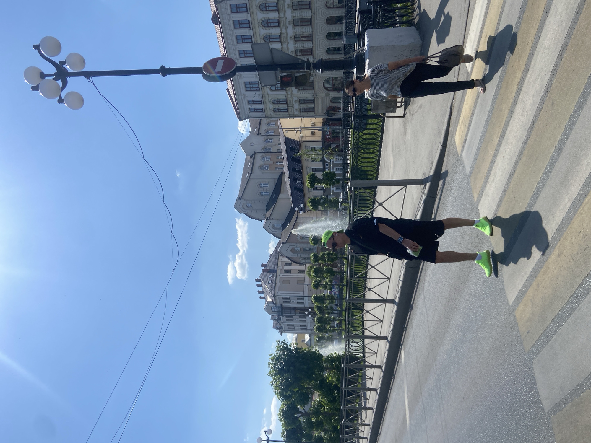 The image shows a street scene in Kazan, Russia, with two people crossing a zebra crossing. One person is wearing black shorts and a black shirt with bright green sneakers and a green hat. The other person is wearing dark pants and a light-colored striped shirt. A fence separates the sidewalk from a fountain with a row of trees. Buildings are visible in the background, along with a lamppost and a traffic sign. The sky is blue with some clouds. The image is taken during the day with clear lighting.