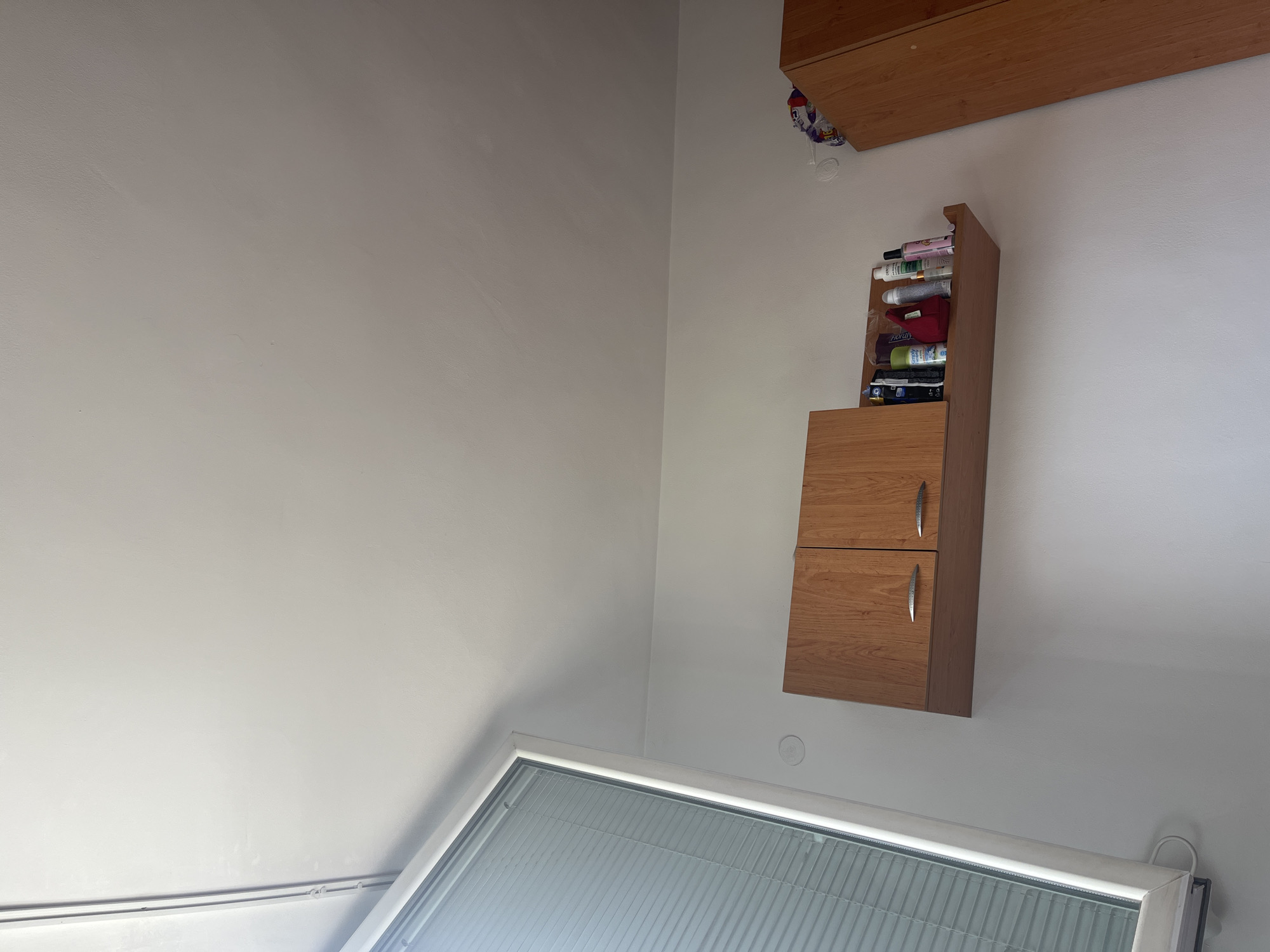 This media file captures an interior scene, likely a room, from a high-angle perspective looking upwards towards a wall and ceiling. The walls are painted a uniform light grey or off-white color.
On the right side of the frame, a light brown wooden cabinet system is mounted on the wall. This system comprises at least two sections: a lower cabinet with two doors, each fitted with a curved silver handle, and an open upper shelf area. The open shelf contains various items, primarily bottles, tubes, and spray cans, suggesting toiletries, cosmetics, or household products. Visible brand names or labels on these items include "POND'S" (partially visible), "Floralys," "Simply" (partially visible), and "GARNIER" (partially visible). Above this unit, another long, narrow wooden shelf or cabinet is visible along the top edge of the frame. A colorful, possibly plastic, item or bag hangs from or near this upper shelf.
In the lower portion of the image, a large skylight window with horizontal blinds is prominent. The window has a white frame and appears to be letting in natural light, indicating it is daytime. A small, white, circular fixture, possibly a smoke detector or light base, is visible on the wall below the wooden cabinet. No people or specific activities are depicted. The scene presents a static view of a functional room space.