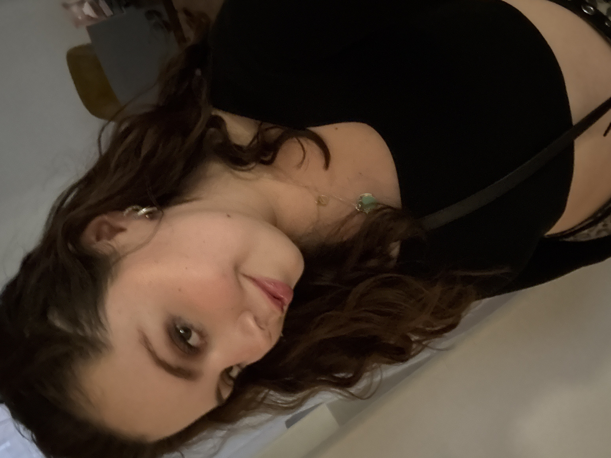 A woman with shoulder-length, curly brown hair is shown from the chest up in an indoor setting. She is wearing a black top with a strap of some kind crossing over her shoulder. She has light skin, and the lighting is somewhat dim, casting shadows on her face and hair. She is wearing small earrings. A necklace with a green pendant is visible. The background is indistinct, but appears to include a wall.