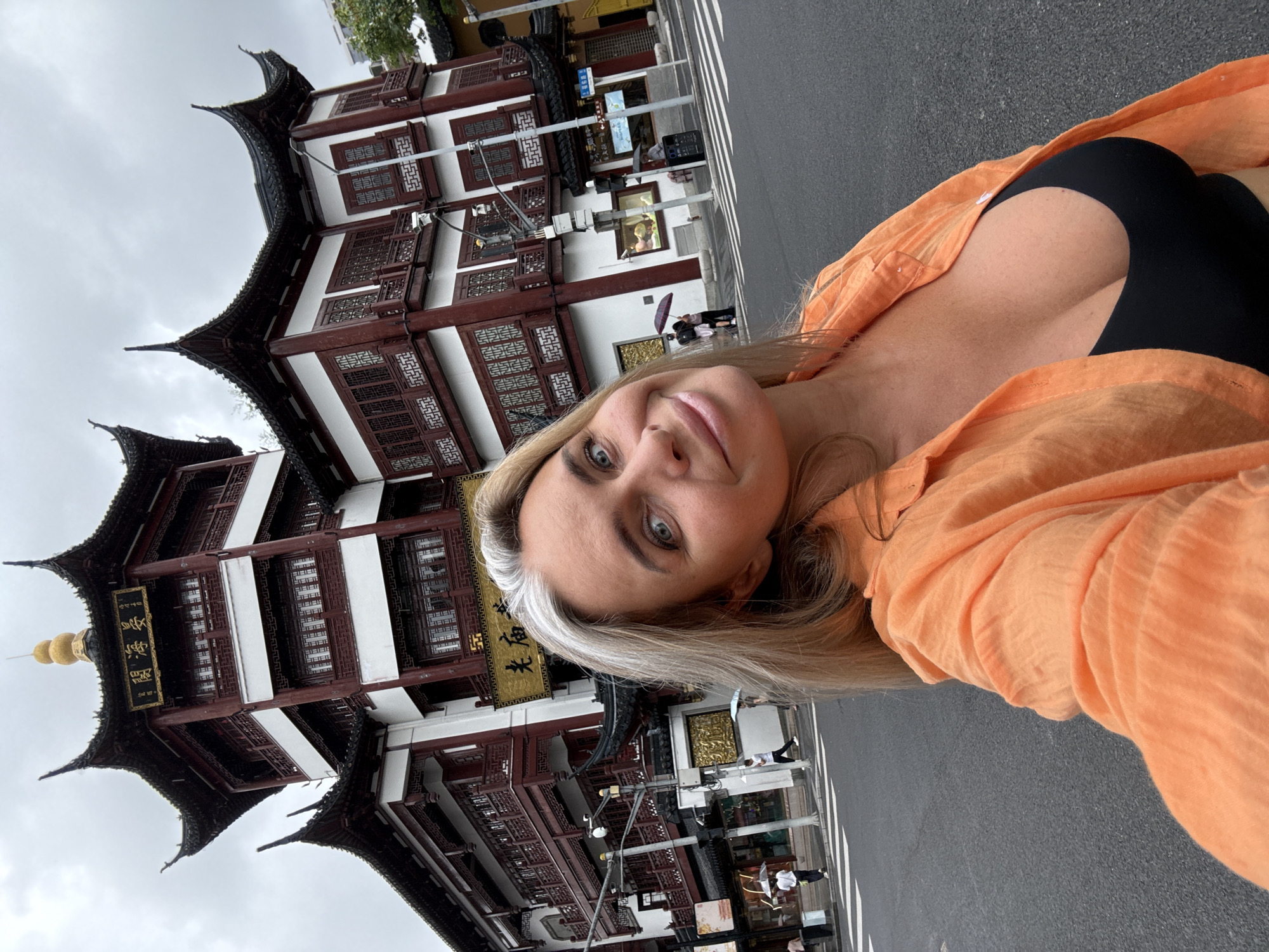 A young Caucasian woman with blonde hair and blue eyes takes a selfie in front of a traditional Chinese building. She is wearing an orange unbuttoned shirt over a black top. The building behind her is red and white with multiple stories and a tiered, curved roof, typical of historical Chinese architecture. The building has gold lettering on its facade. The scene appears to be a street in Shanghai, China, possibly in a historic district like the Yu Garden area, given the architectural style. The sky is overcast. There are other people in the background walking with umbrellas, suggesting it might be raining. The woman is looking directly at the camera with a slight smile.
