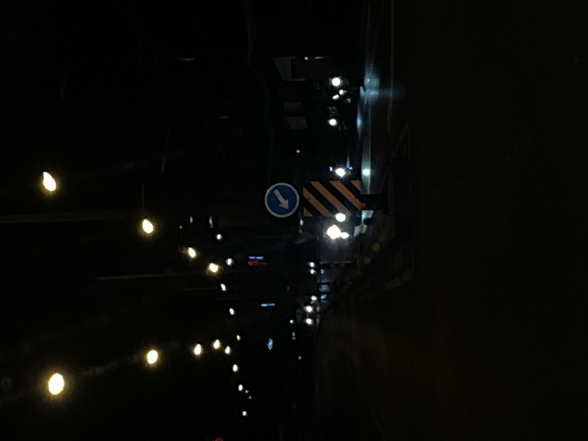 The image shows a street scene at night in Chalong, Thailand. Streetlights illuminate the road, which appears to be wet, reflecting the lights. A blue traffic sign with a white arrow pointing left is visible, mounted on a post with black and yellow diagonal stripes. Several vehicles with their headlights on are also present on the road, with some of them blurred due to motion. The overall atmosphere is dark, with the lighting sources being artificial.
