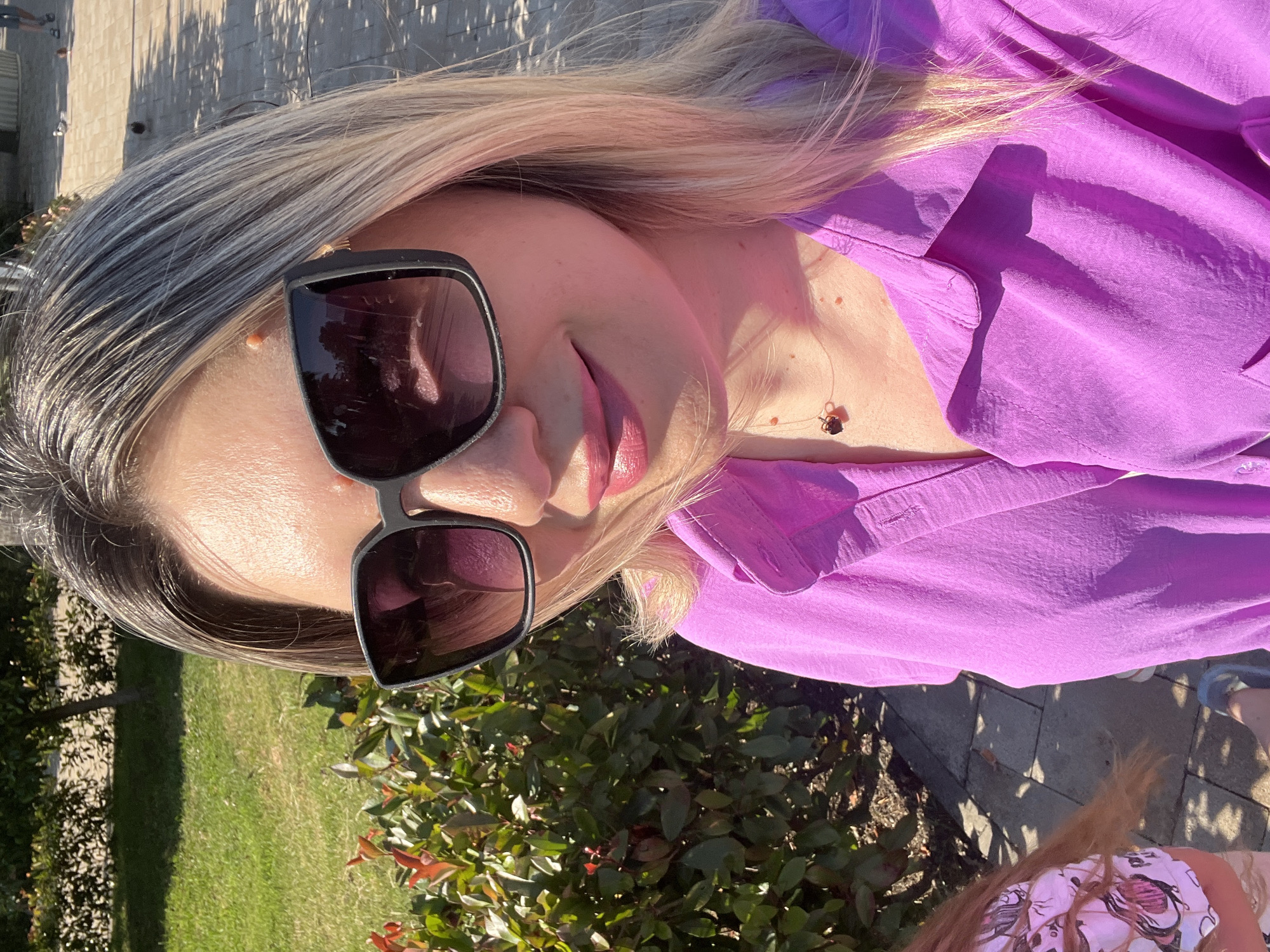 The media file is a close-up, low-angle selfie of a woman outdoors. She has blonde hair with streaks of gray and is wearing large, dark sunglasses. Her lips are painted a glossy pink. She is wearing a vibrant purple shirt. A delicate necklace with a dark pendant is visible on her neck. The setting appears to be a sunny day, indicated by the bright lighting and strong shadows cast on the pavement behind her. Green grass and lush foliage are visible in the foreground and background, suggesting an outdoor, possibly garden or park-like, environment. The overall mood seems relaxed and casual, as if she is enjoying a sunny day outdoors. The time of day appears to be afternoon due to the angle of the sun creating distinct shadows. There is no visible text in the image. The location context is Sochi, Russia.