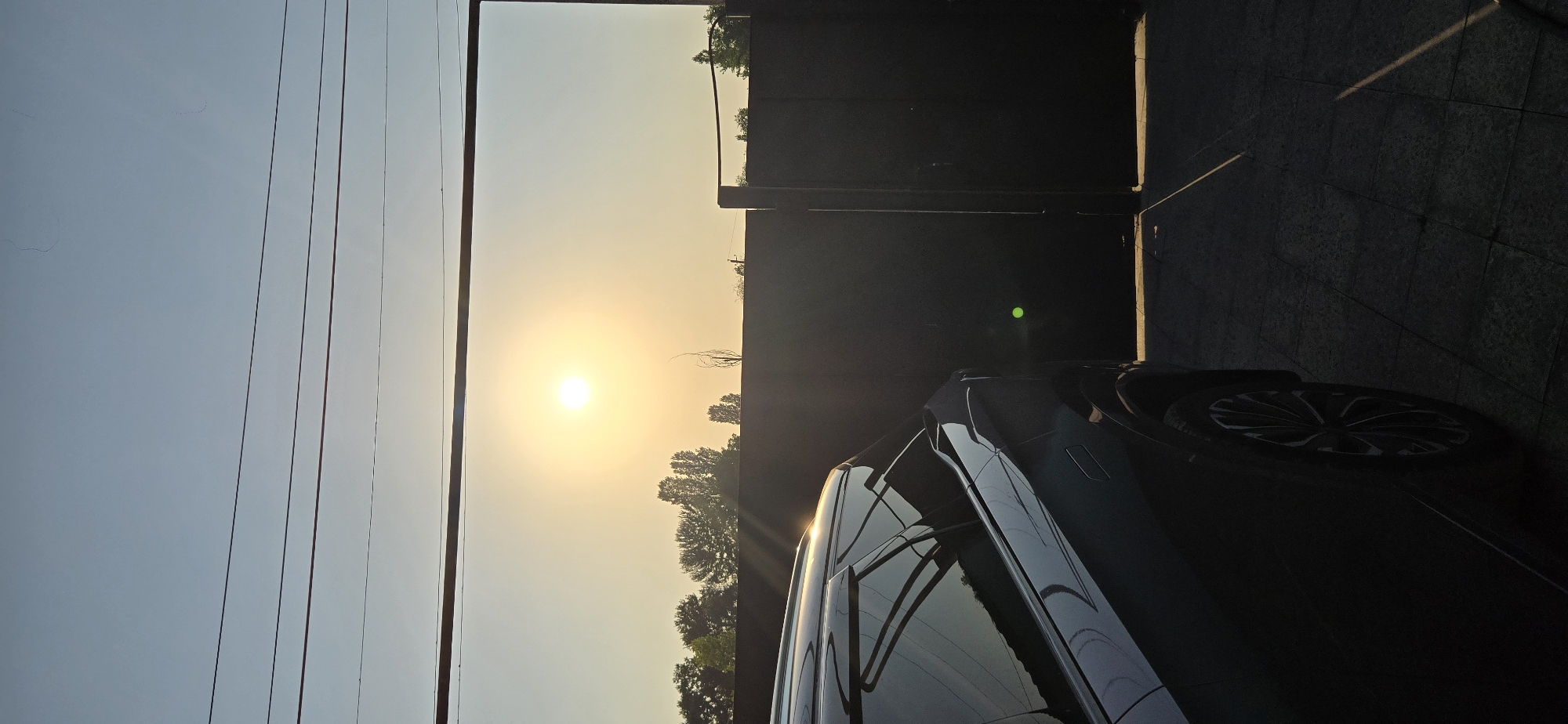 The image depicts a scene in Tel, Azerbaijan, at sunset. A dark-colored SUV is parked in the foreground on a paved surface. The vehicle's windows are reflective, mirroring the sky and trees. Behind the car, a dark, tall gate is visible, partially obscuring the landscape. The sun is low in the sky, casting a warm glow and creating lens flare, appearing as a bright orb in a hazy sky. Silhouetted trees are visible behind a low wall or fence. Overhead, power lines stretch across the frame. The overall atmosphere is tranquil, characteristic of a late afternoon or early evening in a residential or suburban area.
