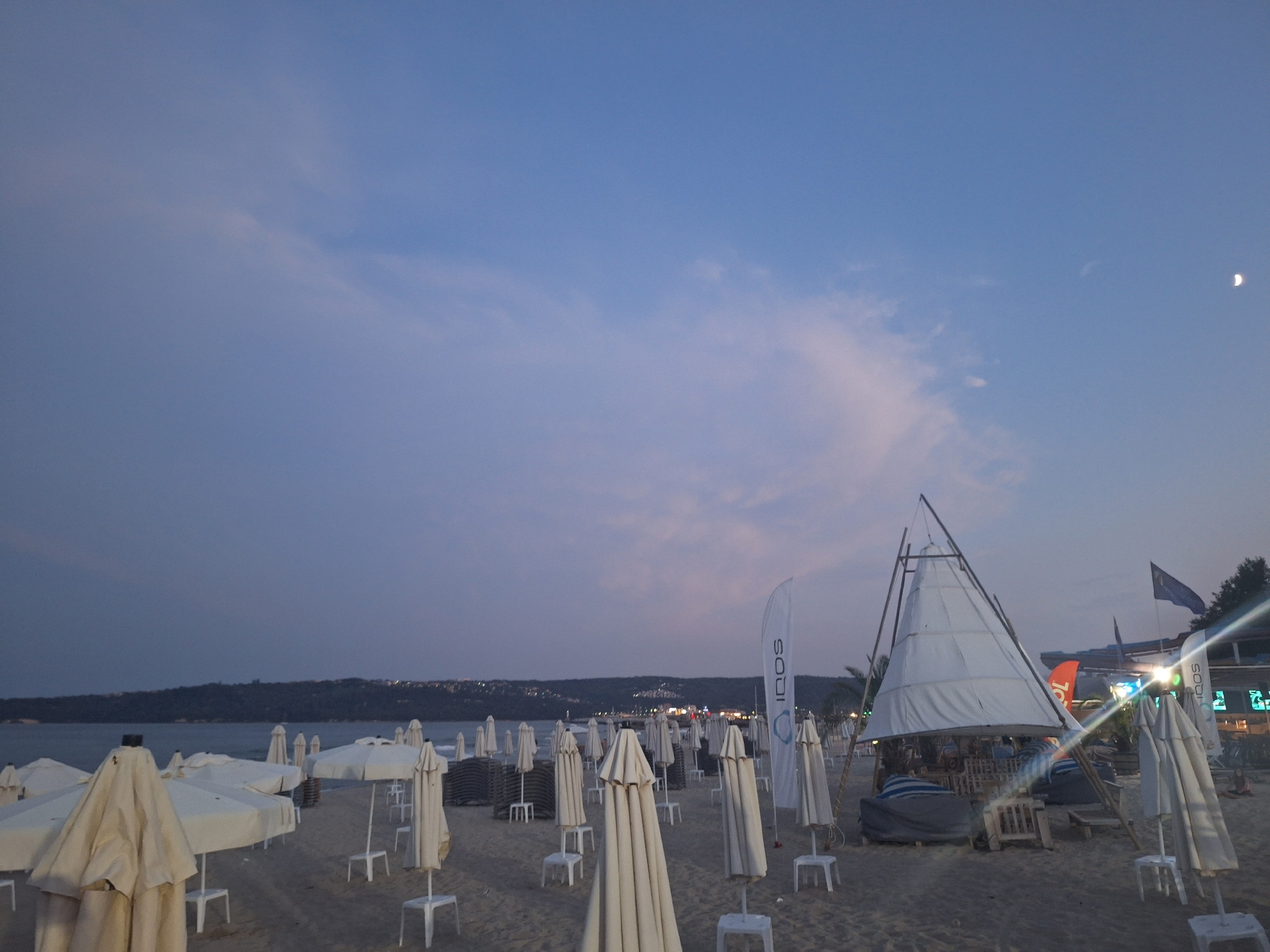The image depicts a beach scene in Varna, Bulgaria, at dusk. Numerous closed beach umbrellas and lounge chairs are spread across the sandy shore. In the background, a body of water is visible, with a distant shoreline marked by hills and scattered lights indicating a town or resort. The sky is a soft blue with wispy clouds, and a crescent moon is faintly visible.
The foreground is dominated by rows of folded white beach umbrellas, each mounted on a white stand. Some umbrellas are in bundles, stacked neatly, while others stand individually. Several small white plastic stools are placed around the umbrellas.
Further back, a distinctive tent-like structure with a white fabric roof supported by wooden poles stands prominently. Adjacent to it, a tall, slender white banner with the text "IQOS" is planted in the sand. Other flags, including a dark blue one, are also visible in the distance.
The lighting suggests it is either early evening or late afternoon, with the light beginning to fade. There are no prominent people visible in the immediate foreground, though a few indistinct figures might be present in the distance near the structures on the shoreline. The overall impression is one of a beach winding down for the day, with facilities prepared for the next.