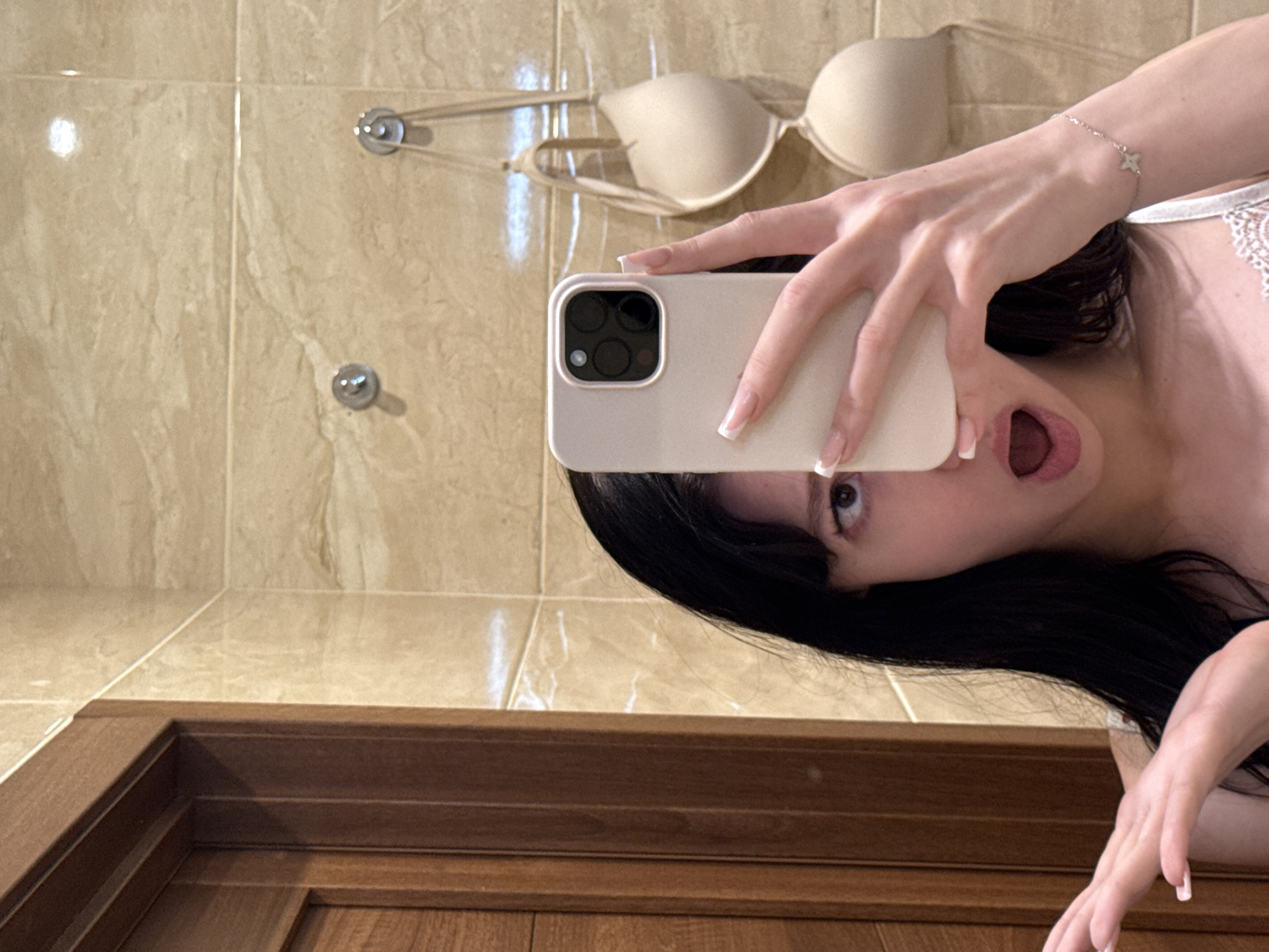 A young woman with long, dark hair and light-colored nail polish is taking a selfie in a bathroom. She is partially obscured by her phone, which she is holding in her right hand. Her mouth is open in an "O" shape, and her left hand is extended outward. In the background, a beige bra is hanging on a hook. The walls are tiled in a beige, marble-like pattern, and there is a wooden door on the left side of the frame. The image appears to have been taken in a residential bathroom, likely in Khimki, Russia, given the context of the request.