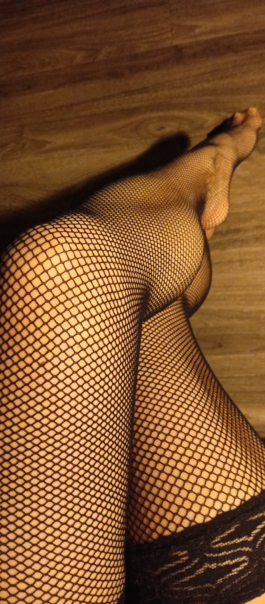 A close-up photograph shows a pair of legs clad in fishnet stockings, crossed at the ankles, resting on a wooden floor. The stockings are dark, and the texture of the fishnet is clearly visible. The skin underneath the stockings is light in color. The top of one stocking has a patterned trim. The background is a wooden floor with a warm, yellowish-brown tone. The lighting casts a shadow of the leg onto the floor. The location is not discernible from the image.