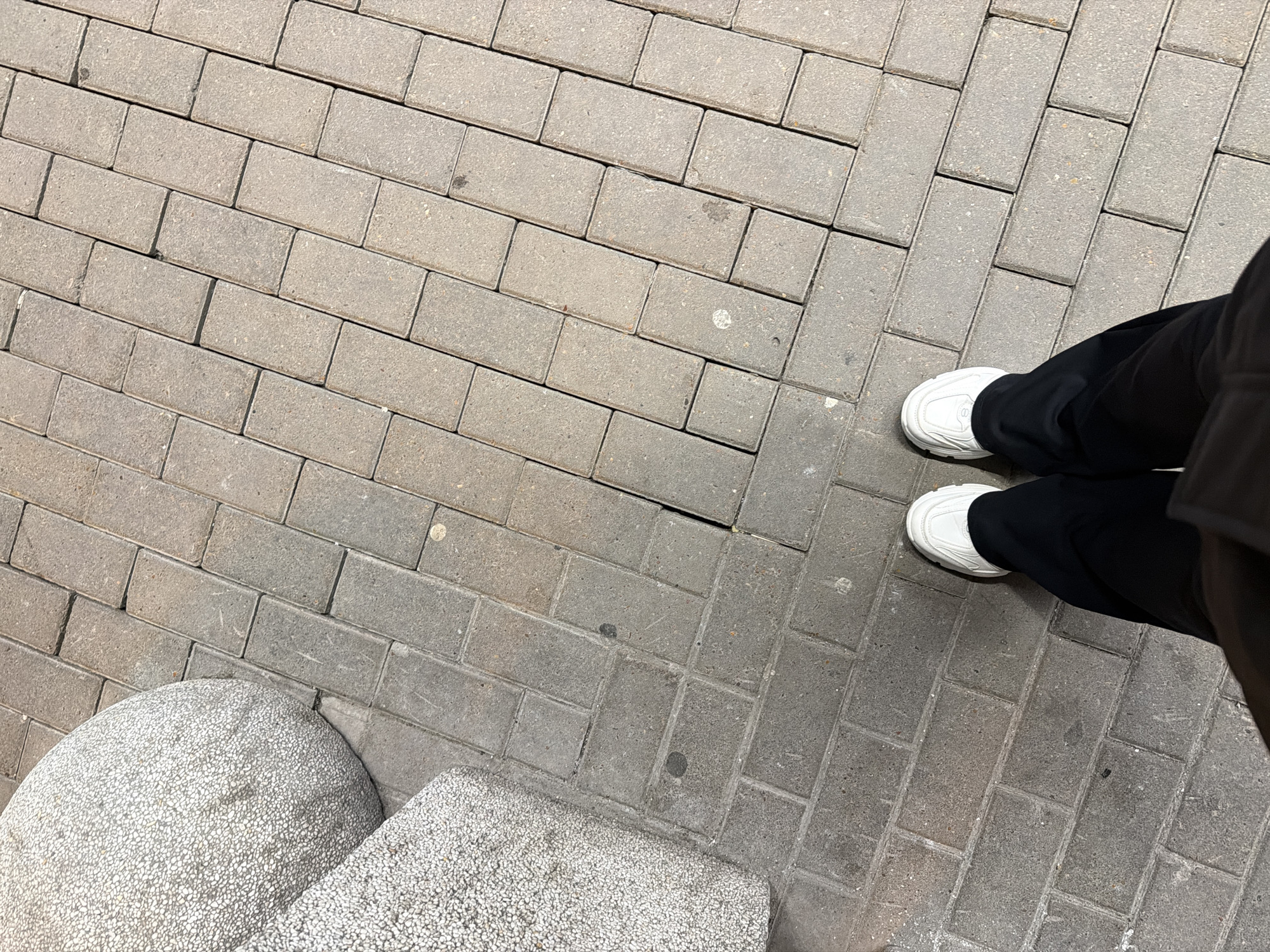 The image shows a section of a paved public area in Ufa, Russia. The paving is constructed from rectangular grey bricks laid in a running bond pattern. To the lower left, part of a speckled stone bench or bollard is visible. In the upper right of the image, a pair of black-clad legs and white shoes are visible. No specific actions or interactions are discernible, other than someone standing on the pavement. The environment appears to be an outdoor space with artificial paving and urban furniture.