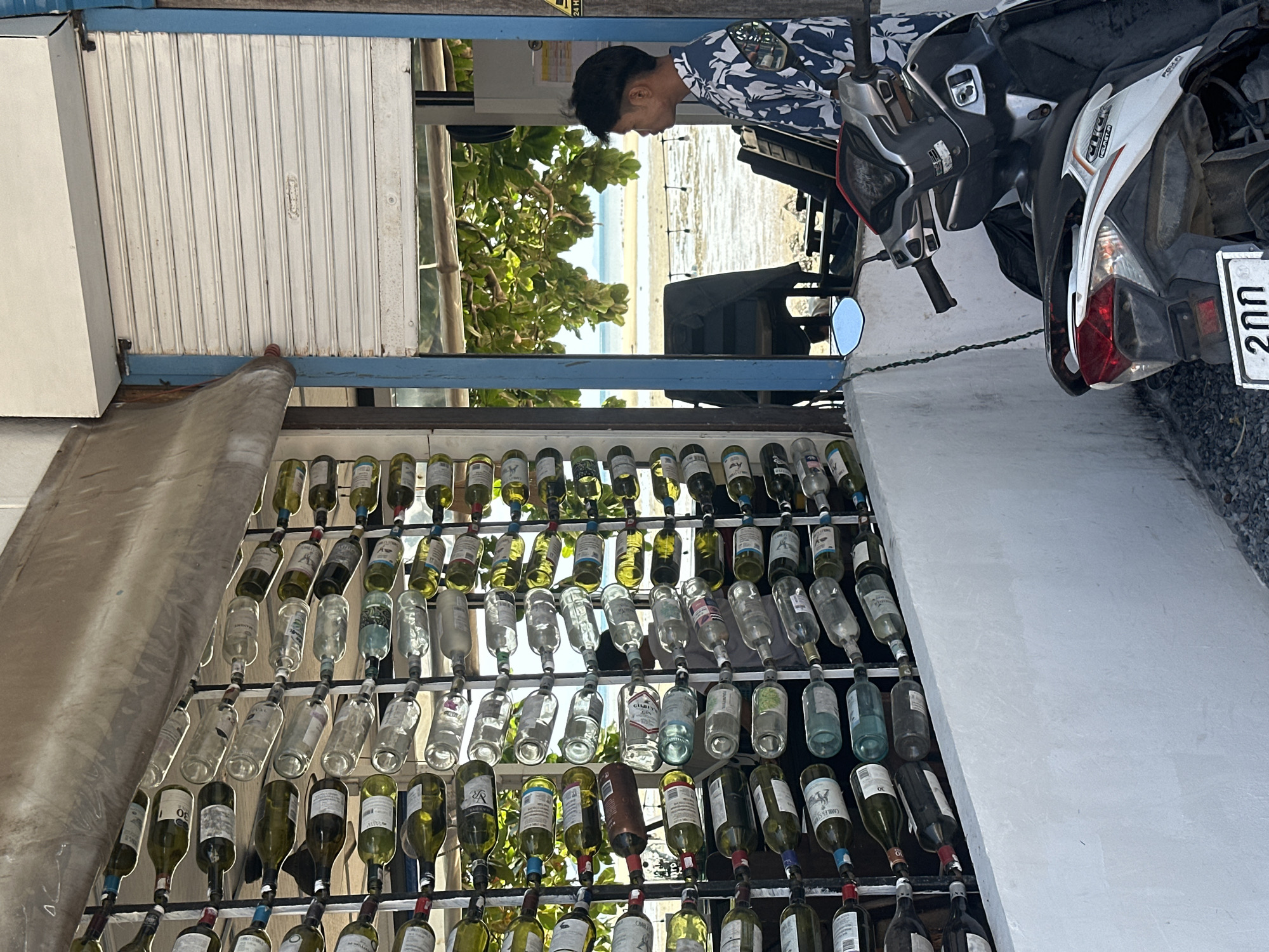 An outdoor view shows a metal wine rack filled with numerous glass bottles, predominantly green and clear, attached to a white building. To the right, a young man wearing a blue and white patterned short-sleeved shirt stands looking out towards a beach scene visible through a blue-framed opening. Behind him, a white roll-up shutter is partially lowered. In the foreground, a modern scooter is parked. The backdrop reveals a sandy beach with calm water and distant trees. The location appears to be a coastal establishment in Koh Phangan, Thailand.