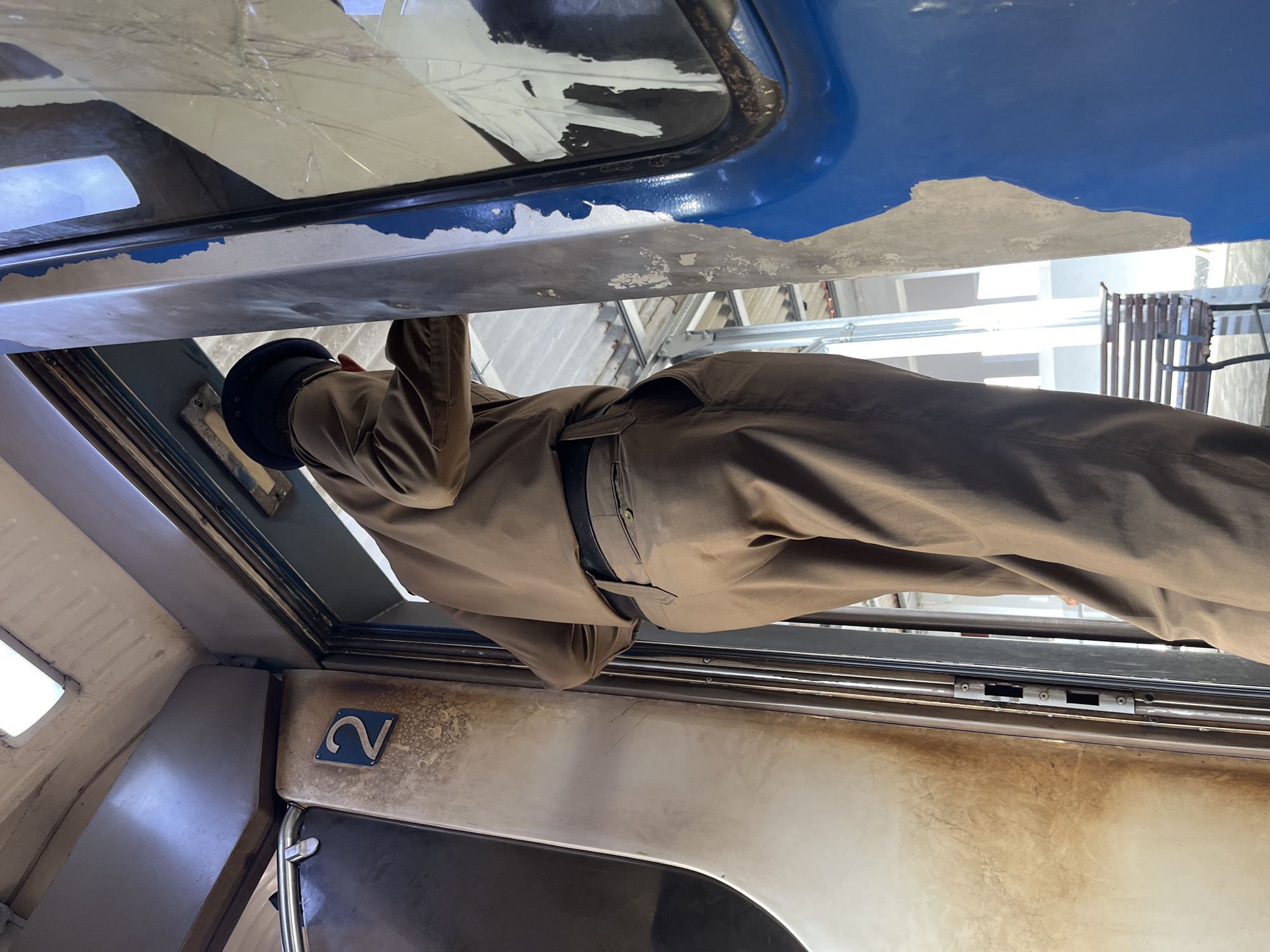 An individual wearing a brown uniform and a hat stands in the doorway of a train carriage, with their back to the camera. The carriage interior is visible, with a "2" marked on the wall. The exterior of the carriage is blue with peeling paint. In the background, a bench is visible on a platform. The scene appears to be a train station in Ambalangoda, Sri Lanka.