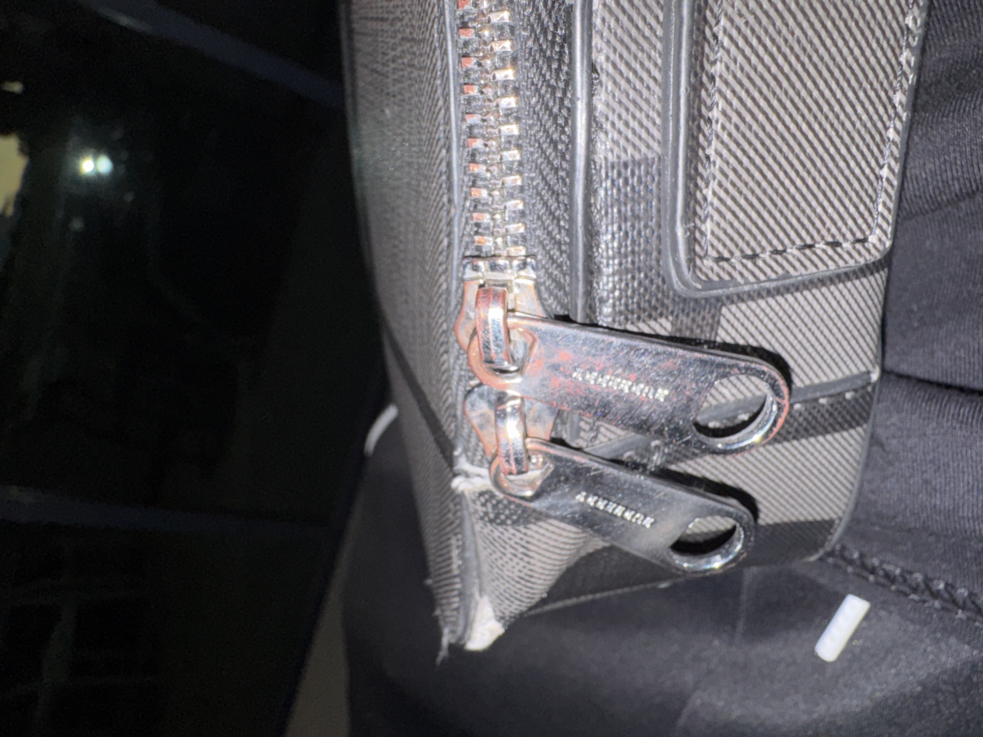 The image presents a close-up view of a metallic zipper and two zipper pulls on an object, likely a bag or backpack. The fabric surrounding the zipper features a distinct striped or plaid pattern in shades of grey and black. The metallic zipper pulls show signs of wear, including scratches and areas of reddish-brown discoloration, possibly tarnish. Fragmented text, appearing to spell "BURBERRY," is engraved on both zipper pulls. Below the zipper, the fabric is visibly frayed and damaged, with white threads exposed. The background is dark, blurry, and out of focus, suggesting a low-light indoor setting. A small, white, cylindrical object is resting on dark fabric to the right of the zipper pulls. No people are visible, and no specific location cues for Yerevan, Armenia, are discernible within the frame. The overall impression is of a well-used item.
