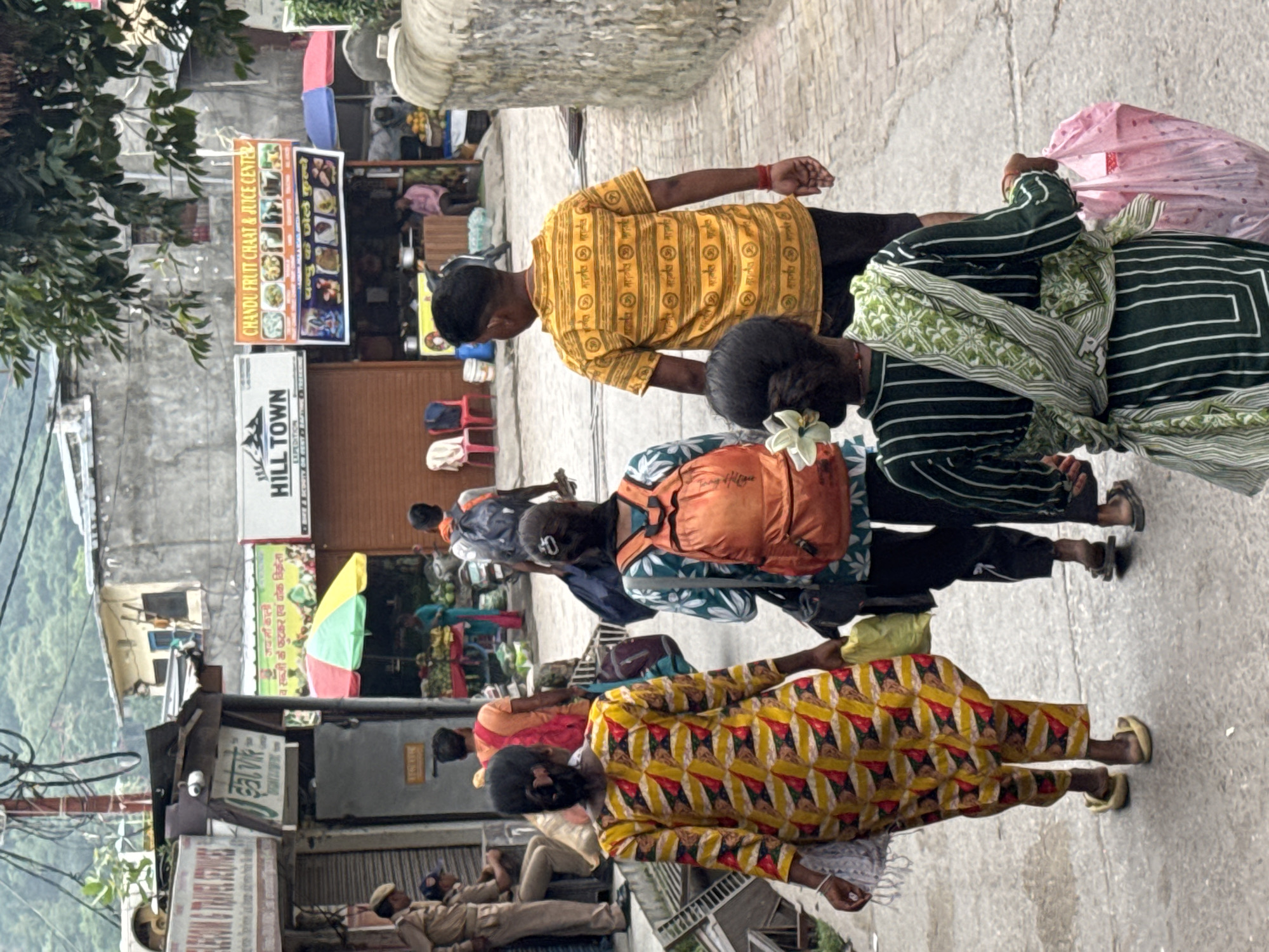 The image depicts a street scene in Rishikesh, India, with several people walking along a paved walkway. In the foreground, a woman in a yellow and red patterned outfit walks away from the camera, carrying a bag. To her right, another woman with an orange backpack walks in the same direction. Behind them, a person in a yellow, horizontally striped shirt with Sanskrit script also walks away from the camera. Several shops are visible on either side of the street, including a "CHANDU FRUIT CHAAT & JUICE CENTER" with an orange banner and colorful umbrellas. In the background, buildings with concrete walls and corrugated metal roofs are visible, with foliage and overhead wires suggesting an urban environment in a hilly region. Two police officers in uniform are seated on a ledge to the left of the frame. The overall atmosphere is that of a busy marketplace or thoroughfare.