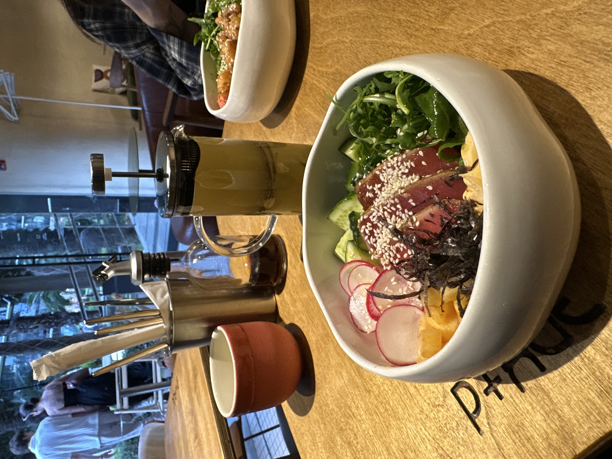 A photograph taken indoors shows a table setting with food and drink in Sochi, Russia.
The table is made of light brown wood, and lettering is carved into the surface. A white, irregular shaped bowl is filled with a colorful salad. Ingredients include leafy greens, sliced cucumbers, slices of red radish, cubes of yellow fruit, and pieces of dark seaweed. Slices of red meat are topped with sesame seeds.
To the left is a glass French press filled with yellow liquid. Next to it is a metal container holding straws and chopsticks, along with a bottle of olive oil. A small red cup sits next to the container. Another white bowl containing food is slightly out of focus in the upper left.
In the background, a window shows people sitting near foliage. A person wearing a plaid shirt is partially visible in the upper left.