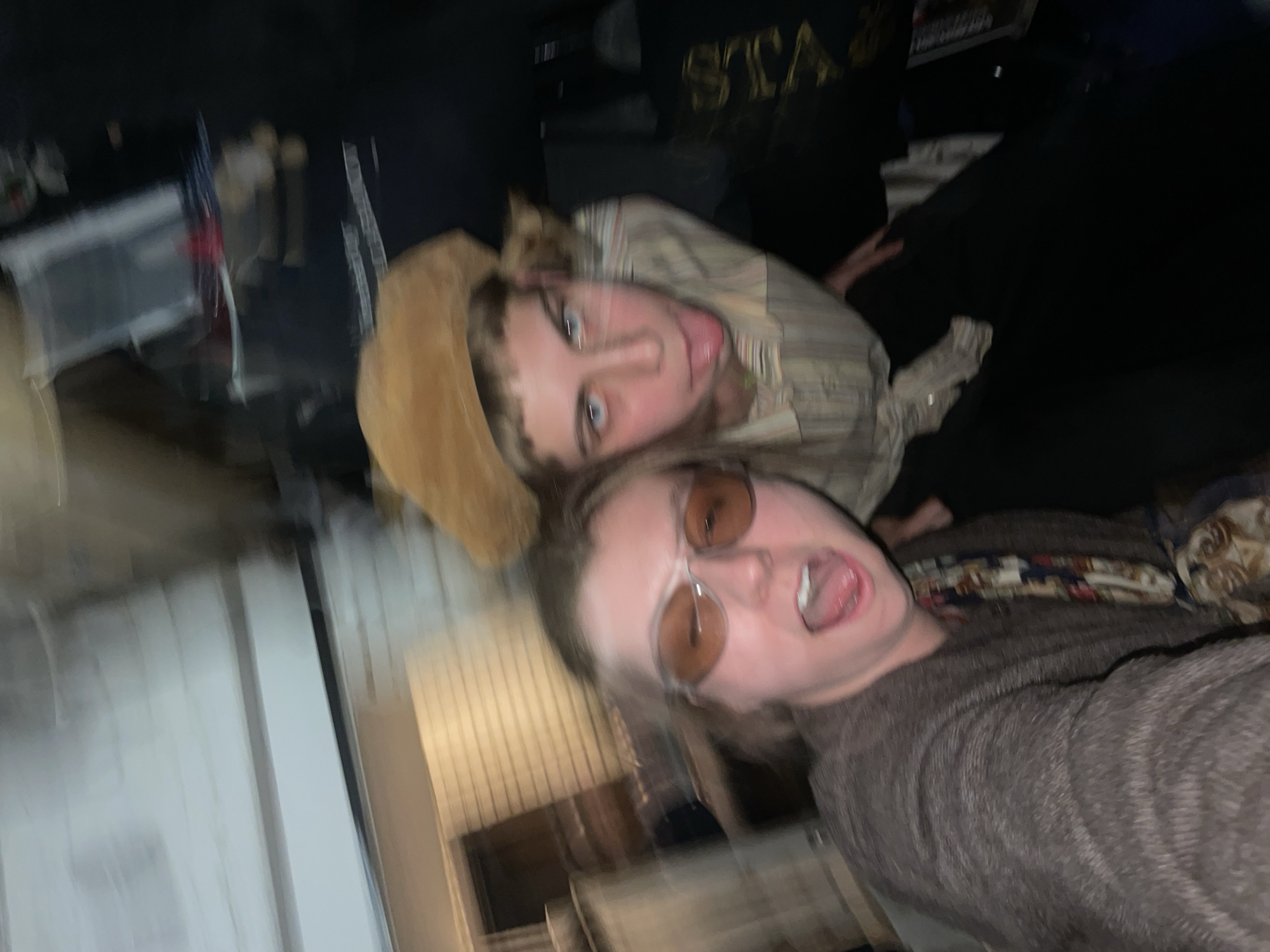 The image shows two individuals, likely indoors, in a blurred and rotated composition. One individual wears a tan cowboy hat and a striped shirt, sticking their tongue out. The other individual wears sunglasses and a dark-colored sweater, also with their mouth open. The background is cluttered with various objects, including what appears to be a flag, a t-shirt with the word "STAR" on it, and storage containers. The location in Moscow, Russia, is not evident from the visual information provided.