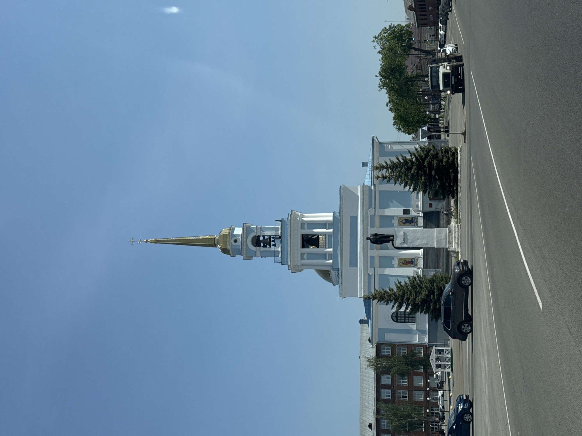 The image depicts the Blagoveshchensky Cathedral in Votkinsk, Russia, viewed from street level. The cathedral, painted in white and pale blue, features a prominent golden spire and multiple levels of architectural detail. A statue of Lenin stands near the base of the cathedral. A two-lane road runs along the cathedral's side. Cars are parked along the roadside. Buildings are visible in the background. The sky is clear and blue.