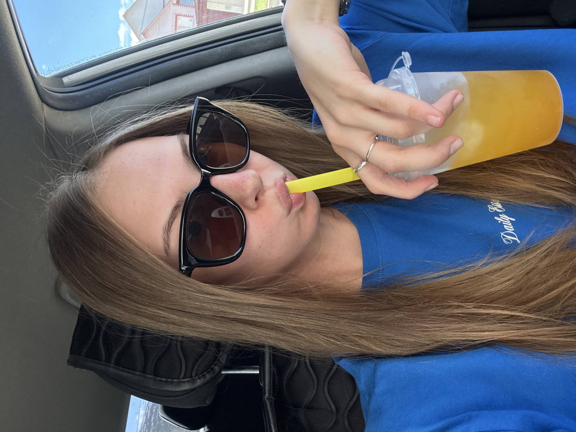 The media file shows a close-up, horizontally oriented selfie of a young woman inside a vehicle. She is wearing a bright blue t-shirt with white text on the chest that reads "Daily". The woman has long, straight brown hair that is parted down the middle and falls over her shoulders. She is wearing large, black-framed sunglasses with brown lenses. Her left hand is holding a clear plastic cup filled with a yellow beverage, and her right hand is holding a yellow straw which she is using to drink from the cup. Her lips are slightly parted around the straw. She is wearing a silver ring on her right ring finger.
The setting appears to be the interior of a car, with a sunroof visible in the upper left corner showing a blue sky with some clouds and a building with a red roof. The interior of the car is dimly lit, suggesting it might be daytime. The woman's expression is neutral. The image appears to have been taken in Perm, Russia, as indicated by the location context.