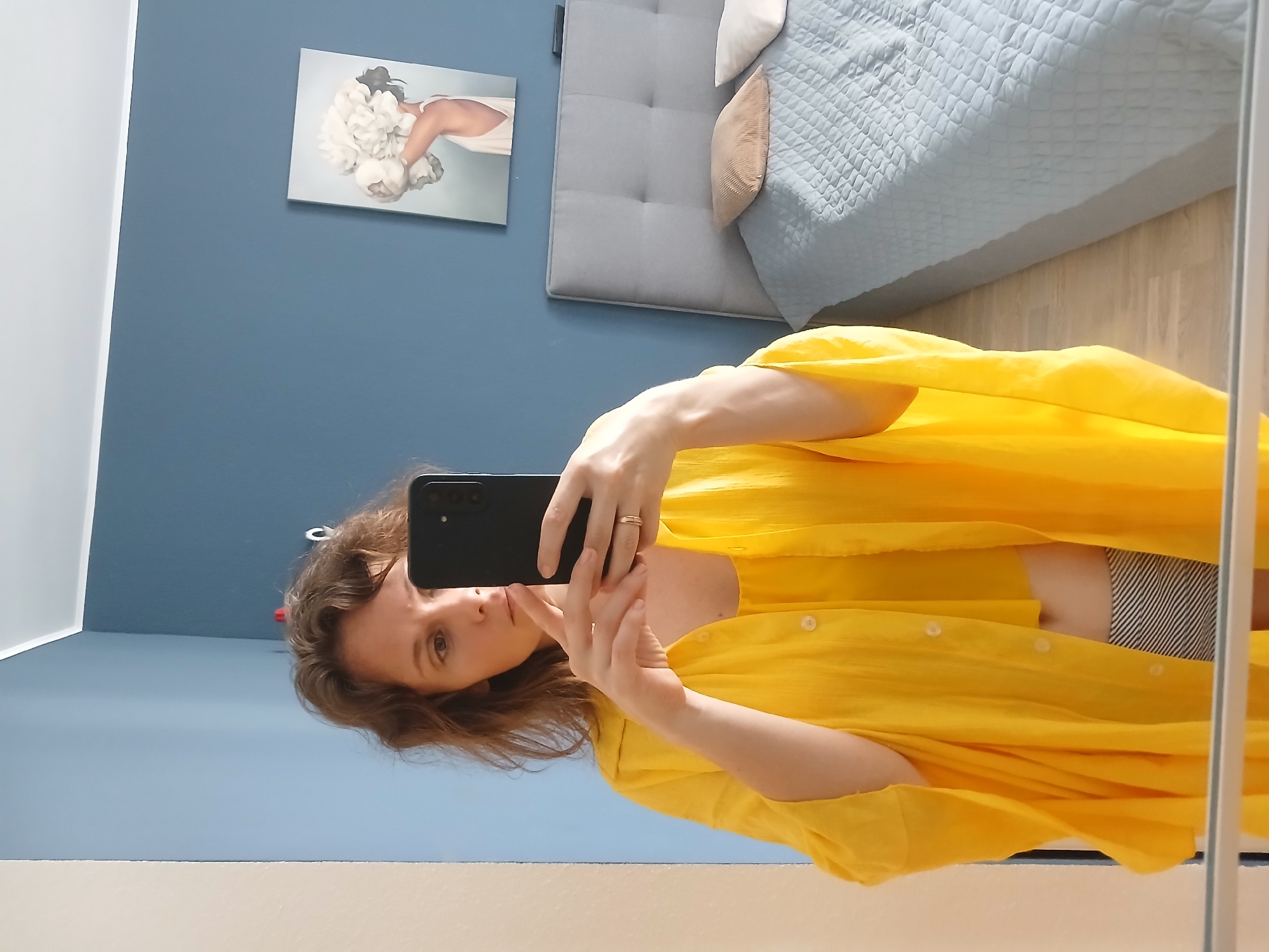In a bedroom with blue walls, a woman stands in front of a mirror, taking a selfie with her phone. She is wearing a yellow top and a matching yellow open shirt over it, with striped shorts. A bed with a grey headboard and light blue bedding is visible in the background, along with a piece of art on the wall depicting a woman holding flowers. The room appears to be a residential interior, likely a bedroom, and there is no visual information to suggest it is located in Yekaterinburg, Russia.