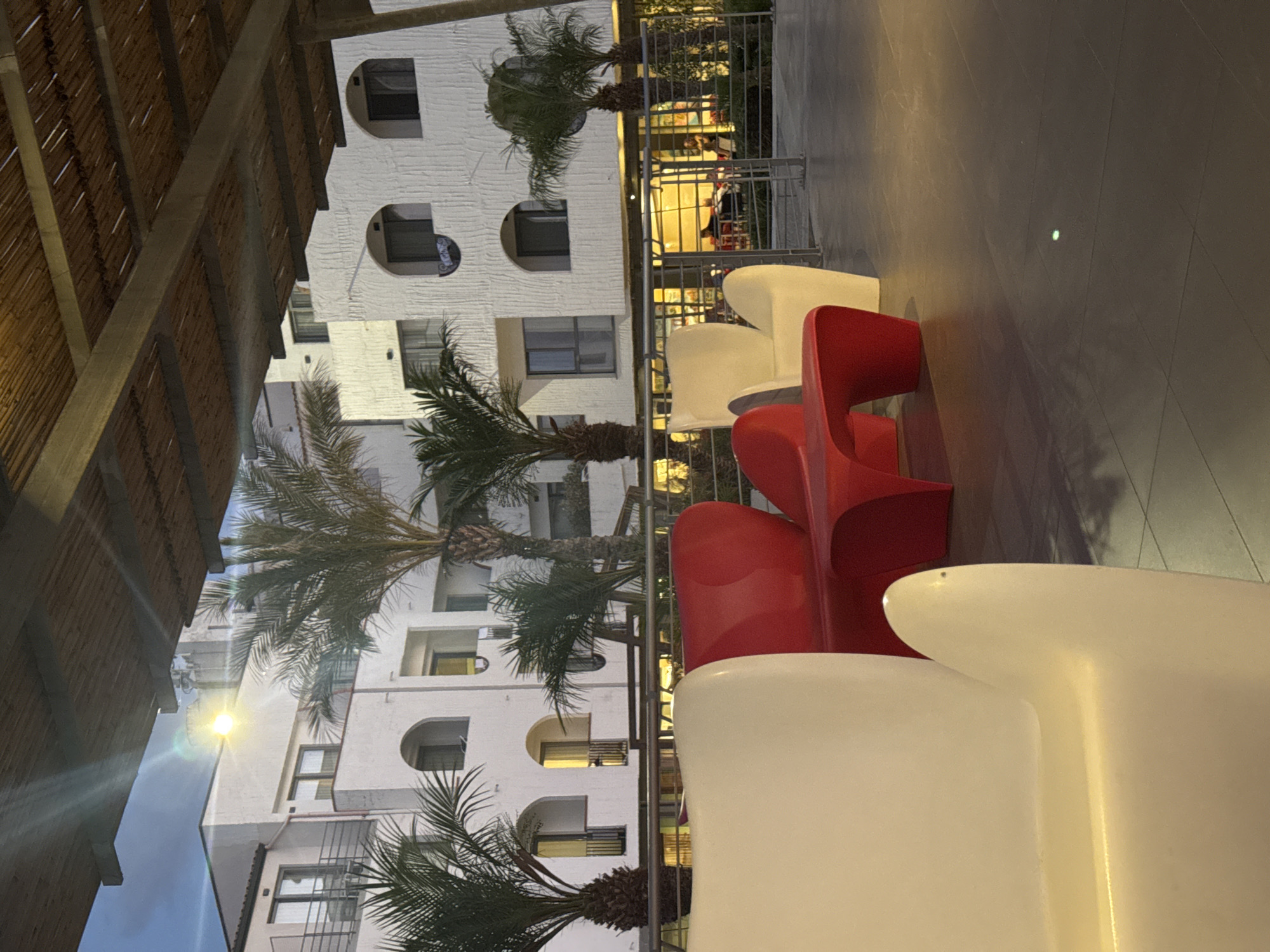 In Isola delle Femmine, Italy, an outdoor seating area is depicted, featuring modern, curved furniture. In the foreground, two white chairs and a red bench with a unique, wave-like design are arranged on a tiled patio. Behind the seating area, palm trees and a multi-story white building with arched windows are visible. A thatched roof structure extends over the seating area, providing shade. The building's facade has a textured finish, and some windows show interior lights. A metal railing separates the patio from a lower level where people are seated at tables, suggesting a restaurant or cafe. The scene is illuminated by artificial lighting, casting shadows and creating a warm ambiance. The sky is visible in the upper left corner, appearing dusky.