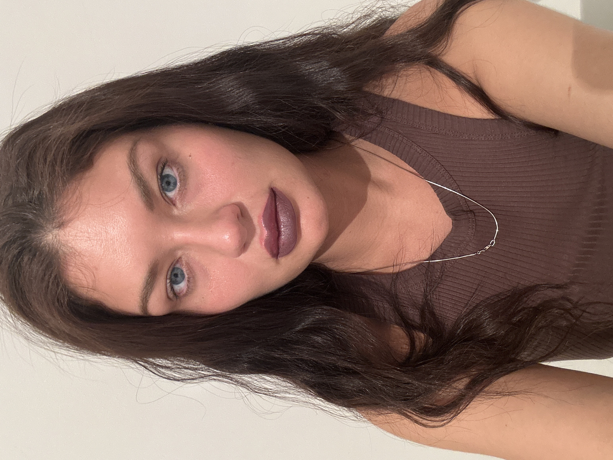 A woman with long dark brown hair and blue eyes is facing the camera. She has a dark mauve lipstick on. She is wearing a brown ribbed tank top and a silver necklace. The background is a plain white wall. The image does not depict Moscow, Russia, or any specific location.