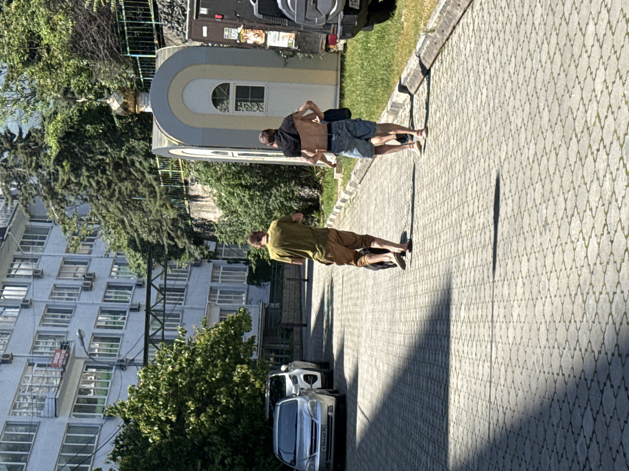 A photo captures a scene in an unidentified city. An apartment building stands to the left, its multiple windows and air conditioning units visible. In the foreground, the walkway is paved with patterned blocks. Two men are seen; one wears a green shirt and khaki shorts, and the other is shirtless, clad in a black shirt and denim shorts. They are positioned close together, possibly interacting or moving an object. A gray building with an arched window is behind them, and a dark vehicle is parked nearby. Two cars are parked alongside the walkway. The photo is rotated, giving the impression that the walkway is vertical rather than horizontal.