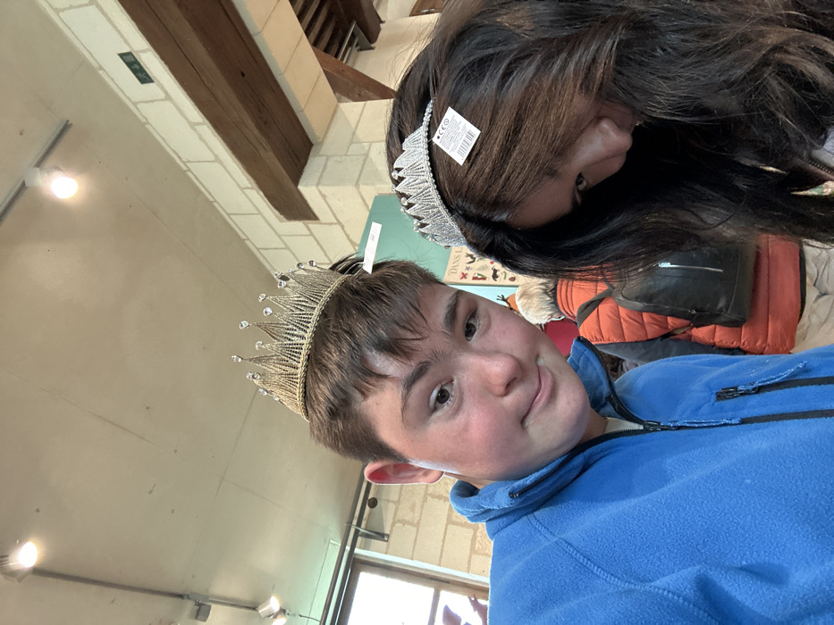 The image depicts two young adults, a male and a female, standing indoors at the Château de Chambord, a royal residence located in the Loire Valley, France, at coordinates 47.616 latitude and 1.518 longitude. The male, wearing a blue fleece jacket, is facing the camera, while the female, with long dark hair, is partially obscured by her hair, looking down. Both are wearing crowns on their heads; the male's crown is gold and the female's is silver. The male has a slight smile on his face. The image was captured in a white-walled room with a wooden beam overhead, and a white tile floor. In the background, a person, possibly a man, wearing an orange jacket and a backpack, is partially visible.