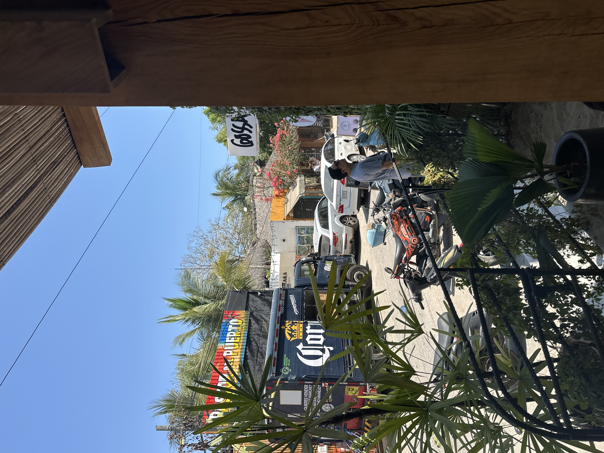 The image depicts a street scene in Puerto Escondido, Mexico. A large black truck with the Corona beer logo and the words "DEL PUERTO" written vertically in colorful letters is parked on the left side of the frame. Cars are parked in front of it. A man in a gray shirt is standing behind the cars. A sign that reads "GOSSA" hangs above a building, indicating the location. A motorcycle is parked nearby. Palm trees and other vegetation are visible throughout the scene. The photo is taken from an elevated position. The sky is clear and blue.