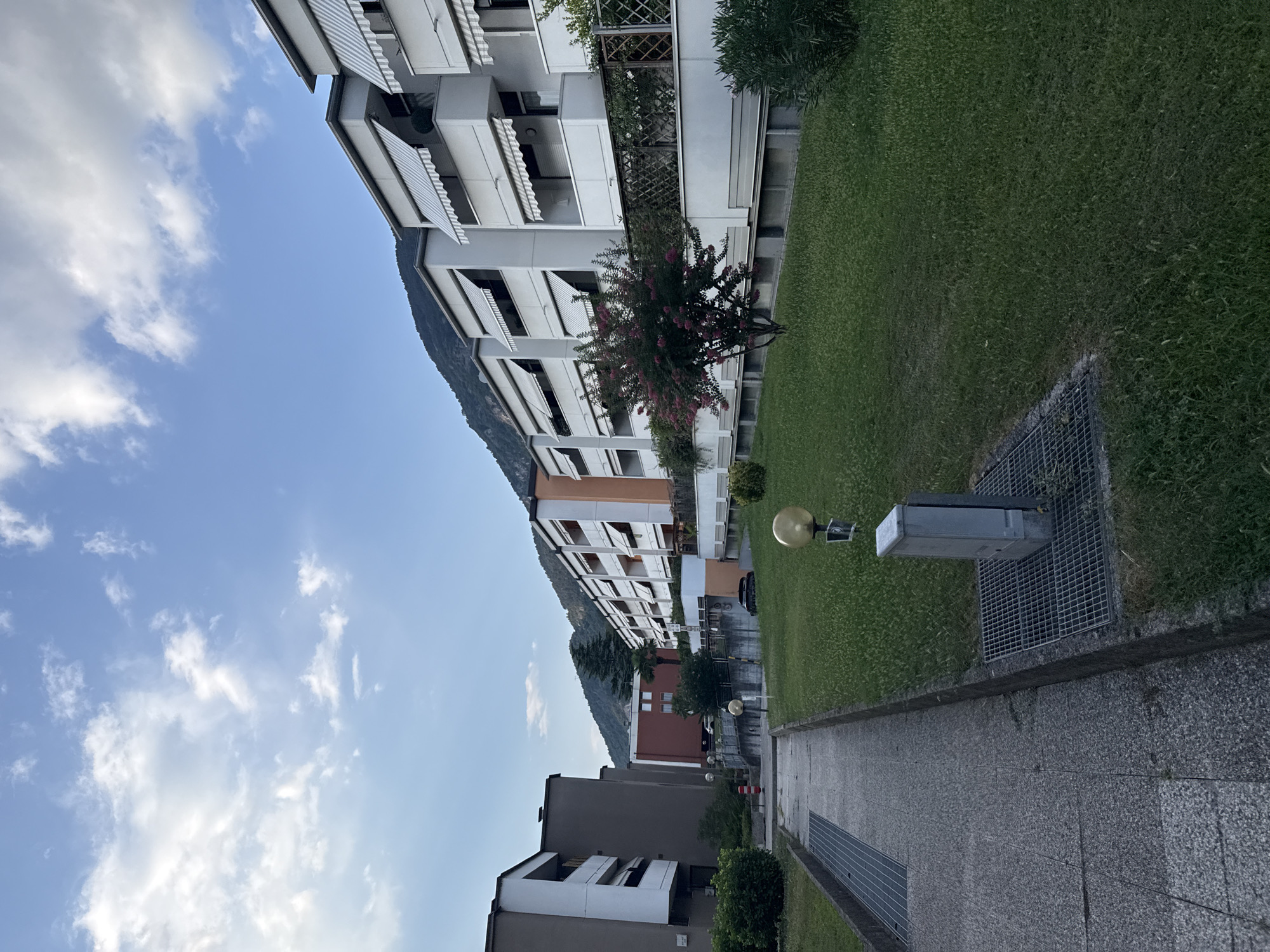 The image shows a street scene in the city of Trento, Italy, featuring modern apartment buildings with white awnings and balconies. A paved walkway runs along the left side of the image, bordered by a grassy area with a metal grate and an electrical box. In the background, mountains rise up, covered in trees and bathed in soft light, indicating it is likely late afternoon or early evening. The sky is a clear blue with scattered white clouds. A person is visible walking on the walkway in the distance. The architecture suggests a residential area, possibly on the outskirts of the city, given the proximity to the natural landscape.