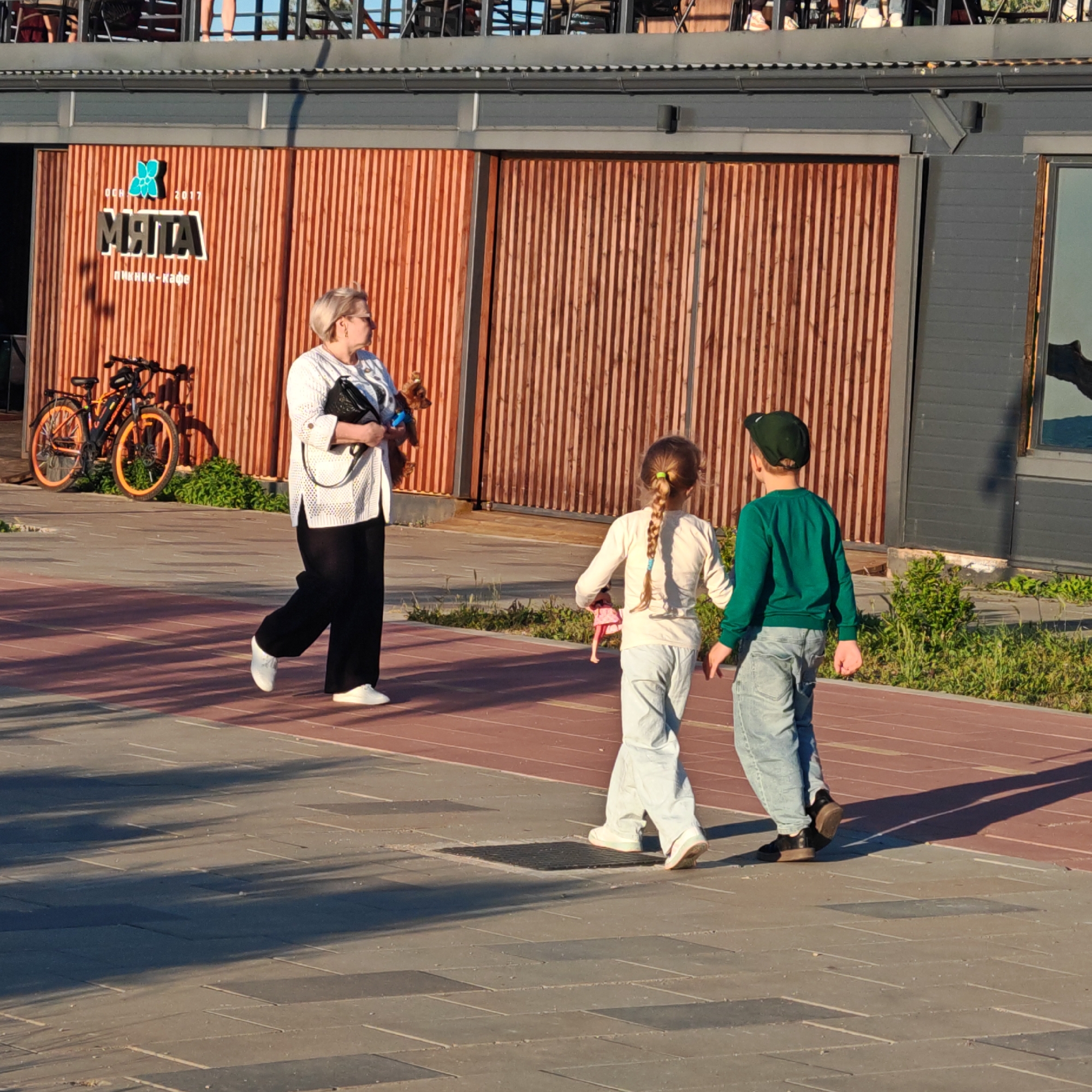 The image depicts an outdoor scene in "городское поселение Щелкино, Ukraine," likely a promenade or public pathway, under the warm, directional light of late afternoon or early evening, evidenced by long shadows.
In the foreground, a paved path with light gray and reddish-brown sections is visible. An adult woman, with short blonde hair and glasses, walks from left to right, wearing a white sweater, black pants, and white shoes. She carries a black bag and holds a leash attached to a small brown dog. Ahead of her, two children walk away from the camera. The girl, with a long braid, wears a light top and jeans and holds a doll. The boy, wearing a dark green cap, a green long-sleeved shirt, and jeans, walks beside her. The overall activity appears to be a casual stroll.
In the background, a building with a modern design features vertical wooden slats on its lower facade and a dark gray upper section with a roof structure, possibly an outdoor seating area. Two bicycles, one prominent with orange accents, are parked against the wooden wall. On the wall, a blue flower-like logo is visible, accompanied by the text "МЯТА" (MYATA), "ПИКНИК-КАФЕ" (PIKNIK-KAFE, meaning "Picnic Cafe"), and "ОСН. 2017" (EST. 2017). Green bushes grow along the base of the building.